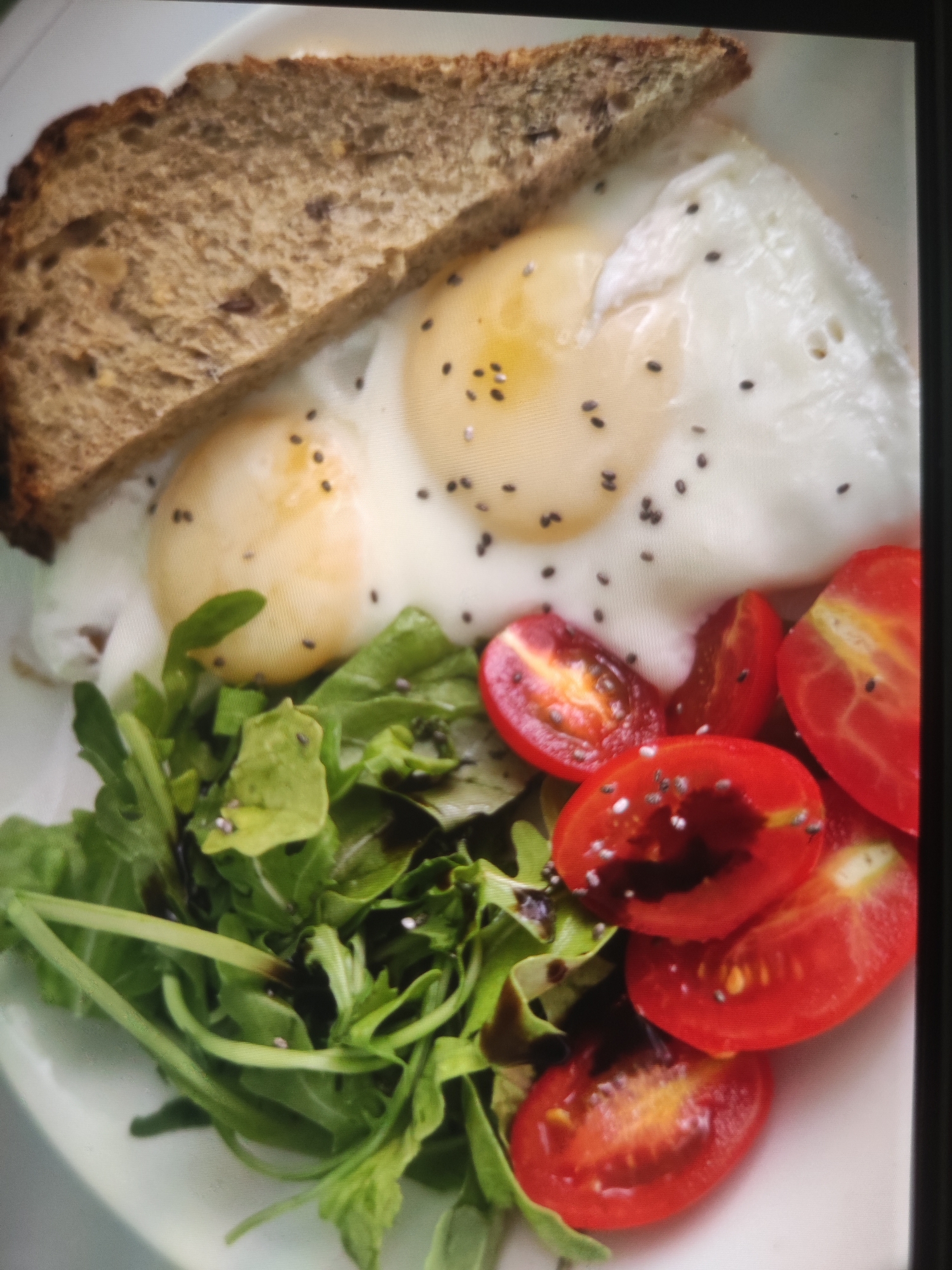 A close-up shot of a plate of food, possibly a breakfast, is captured from a slightly overhead angle. The plate contains two fried eggs with runny yolks, sprinkled with chia seeds. To the left of the eggs is a triangular slice of toasted whole-grain bread. A small salad of green leaves, possibly arugula, and sliced cherry tomatoes is placed in front of the eggs. The salad appears to be dressed with a dark vinaigrette and also has chia seeds scattered on top. The lighting is bright, casting soft shadows. The background is a plain white surface, likely a plate or table. The image does not provide any information about the location in Bökönbaev, Kyrgyzstan.