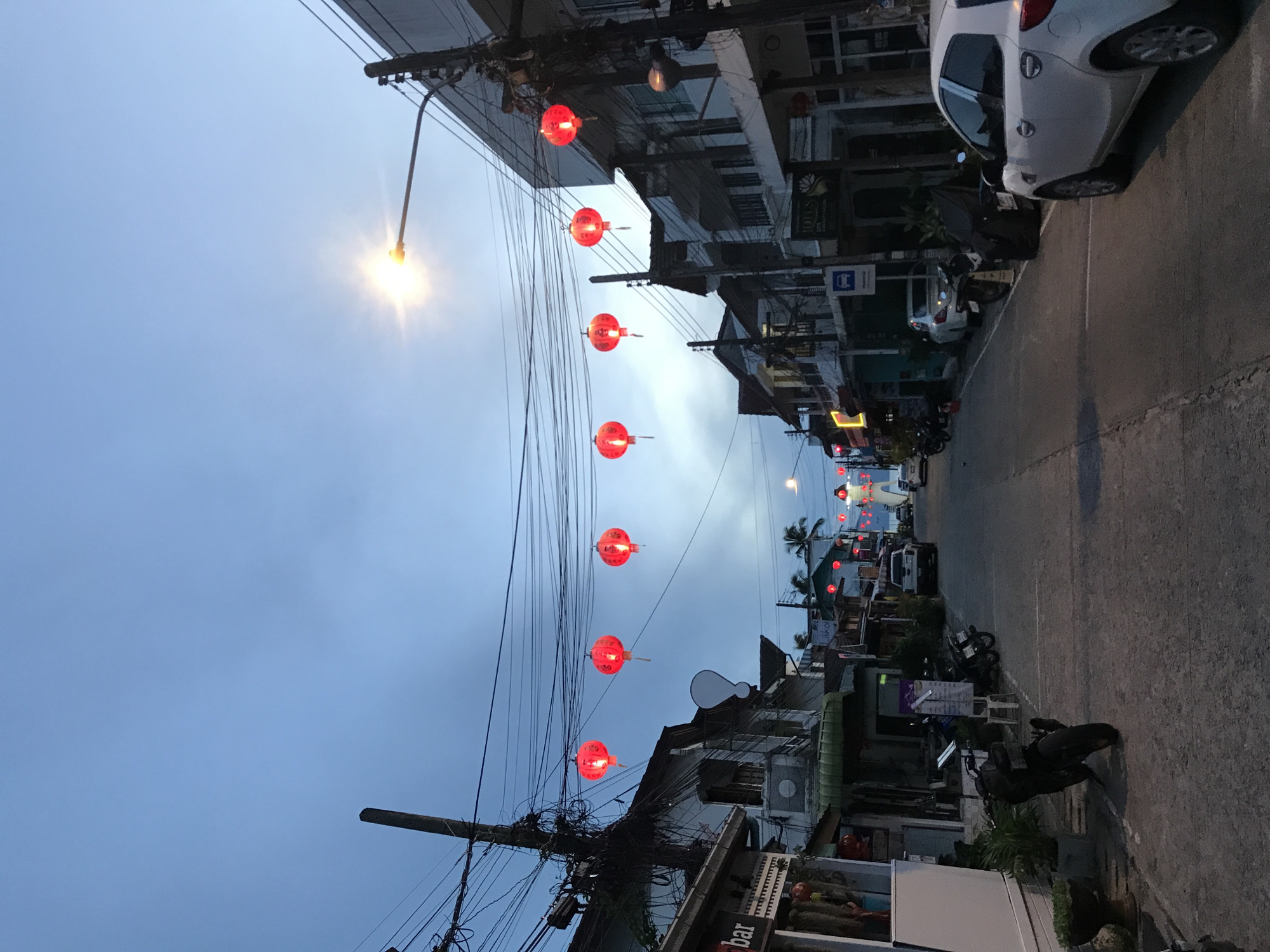A street in Baan Mae Nam, Thailand is captured at dusk. Red lanterns are strung across the street. Buildings and vehicles flank the road. Streetlights illuminate the scene. Overhead power lines are visible.