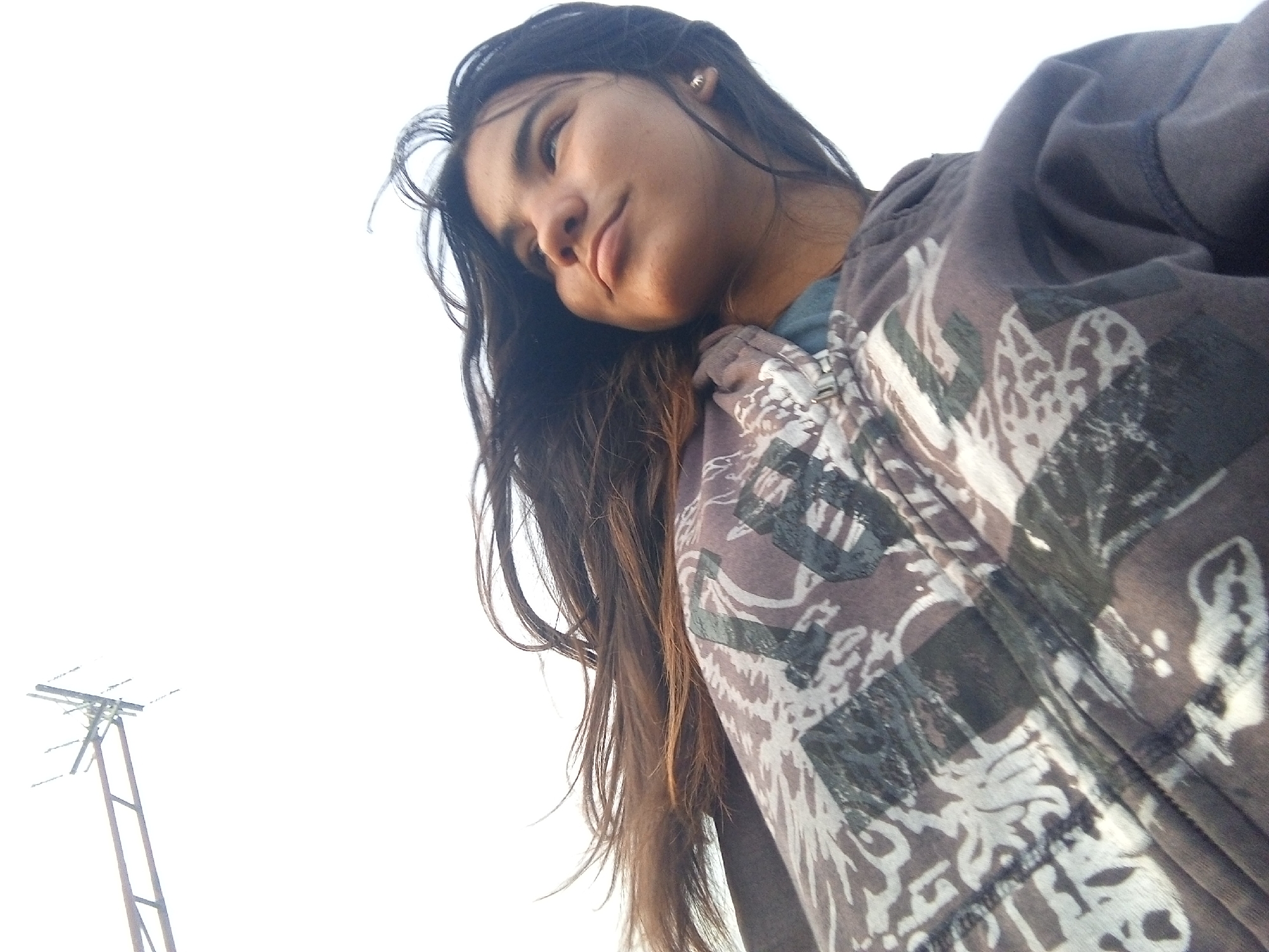 A low-angle shot features a young woman with long, dark hair. She is wearing a patterned zip-up hoodie over a blue top. In the background and to the left, a metal structure resembling a utility pole stands against a bright, white sky. The location is indicated as Altagracia, Venezuela, but there is no other information available about the city from the image.