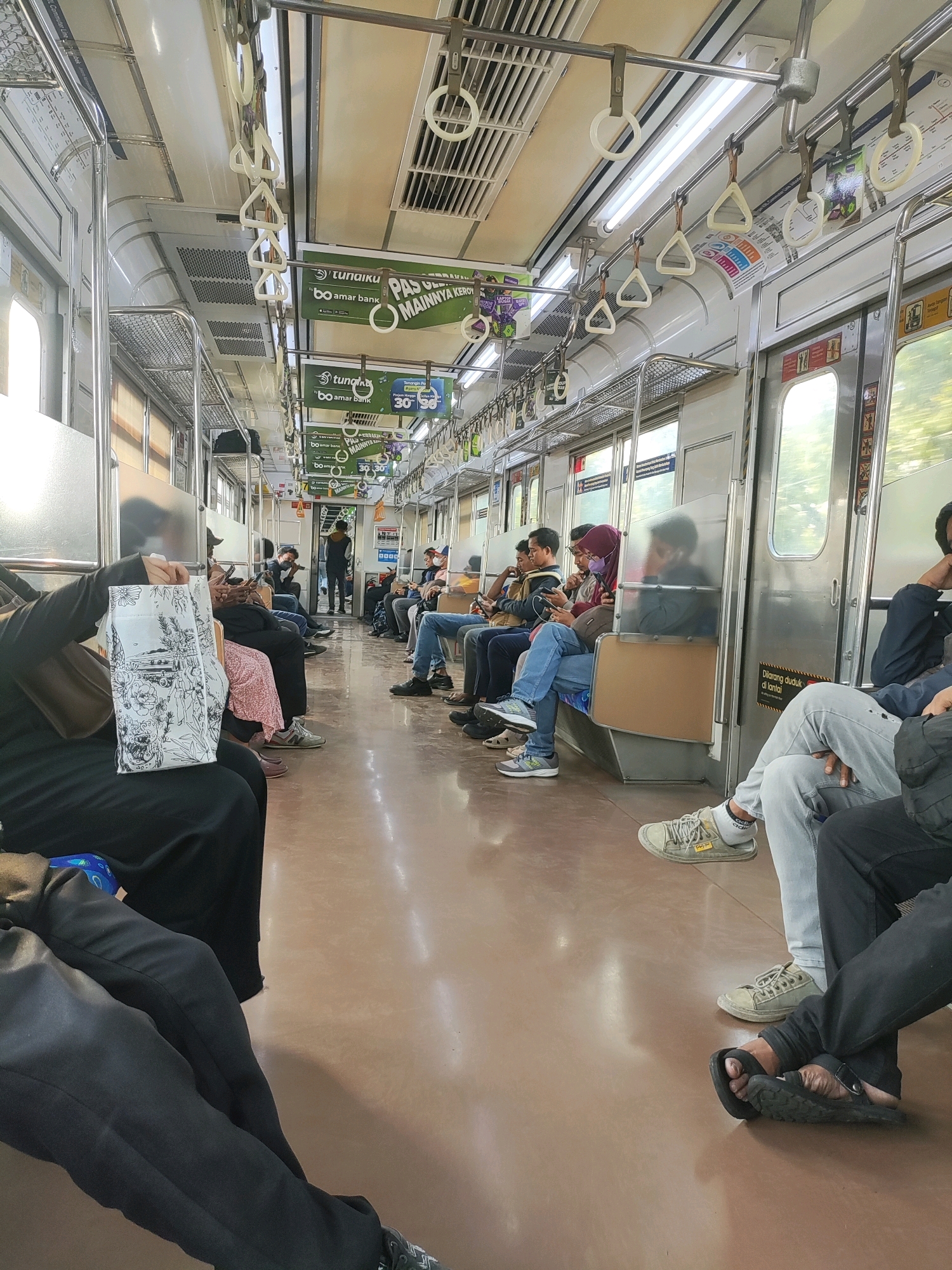 The image depicts the interior of a commuter train carriage in East Jakarta, Indonesia, filled with passengers. The carriage has a light brown floor and a white ceiling with fluorescent lighting and numerous white grab handles for standing passengers. On the right side of the carriage, there are stainless steel doors with windows. Various advertisements are displayed on the walls above the seats, including one that reads "Tunaiku by Amar Bank".
The passengers, a mix of men and women, are mostly seated on the left and right sides of the carriage. Many are looking at their phones or appear to be resting. A woman in the foreground on the left is holding a white tote bag with a black line drawing on it. Another passenger on the right is wearing grey pants and a light-colored sneaker, with their feet crossed. The overall atmosphere is that of a typical public transport commute.