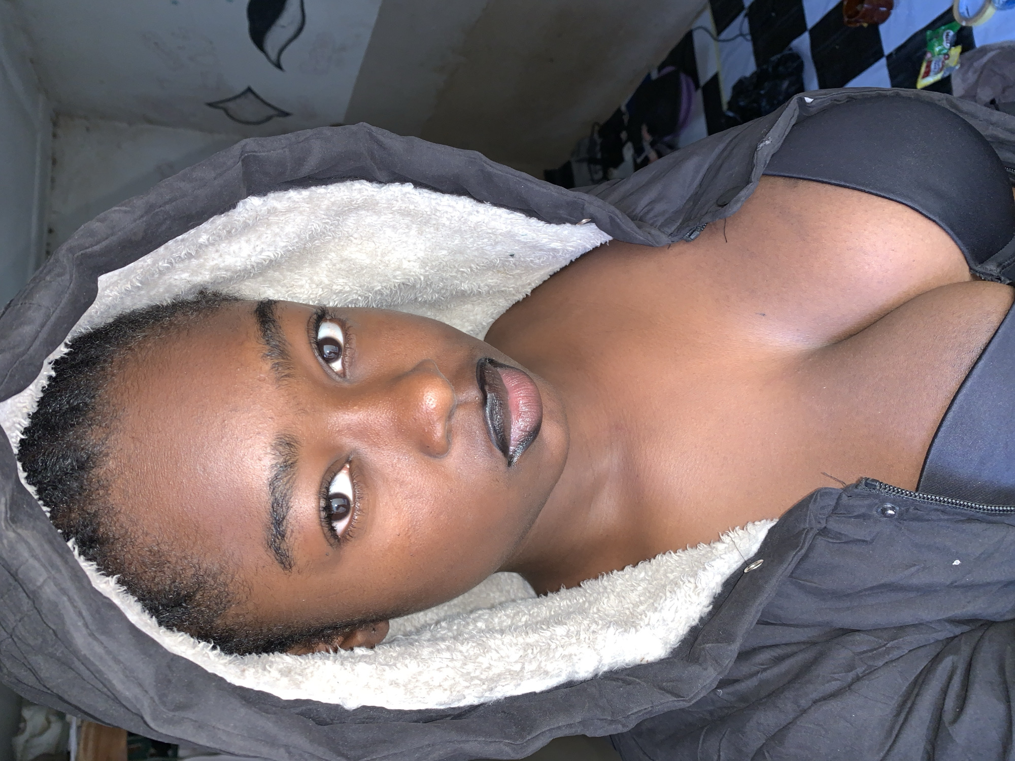 A person with dark skin is lying on a surface, partially covered by a dark jacket with a white, fuzzy lining around the hood. The person's face is visible, and they appear to be wearing dark lipstick. A black bra is visible under the jacket. In the background, there is a black and white checkered surface and some miscellaneous items. The location is identified as Rafin Par, Nigeria.