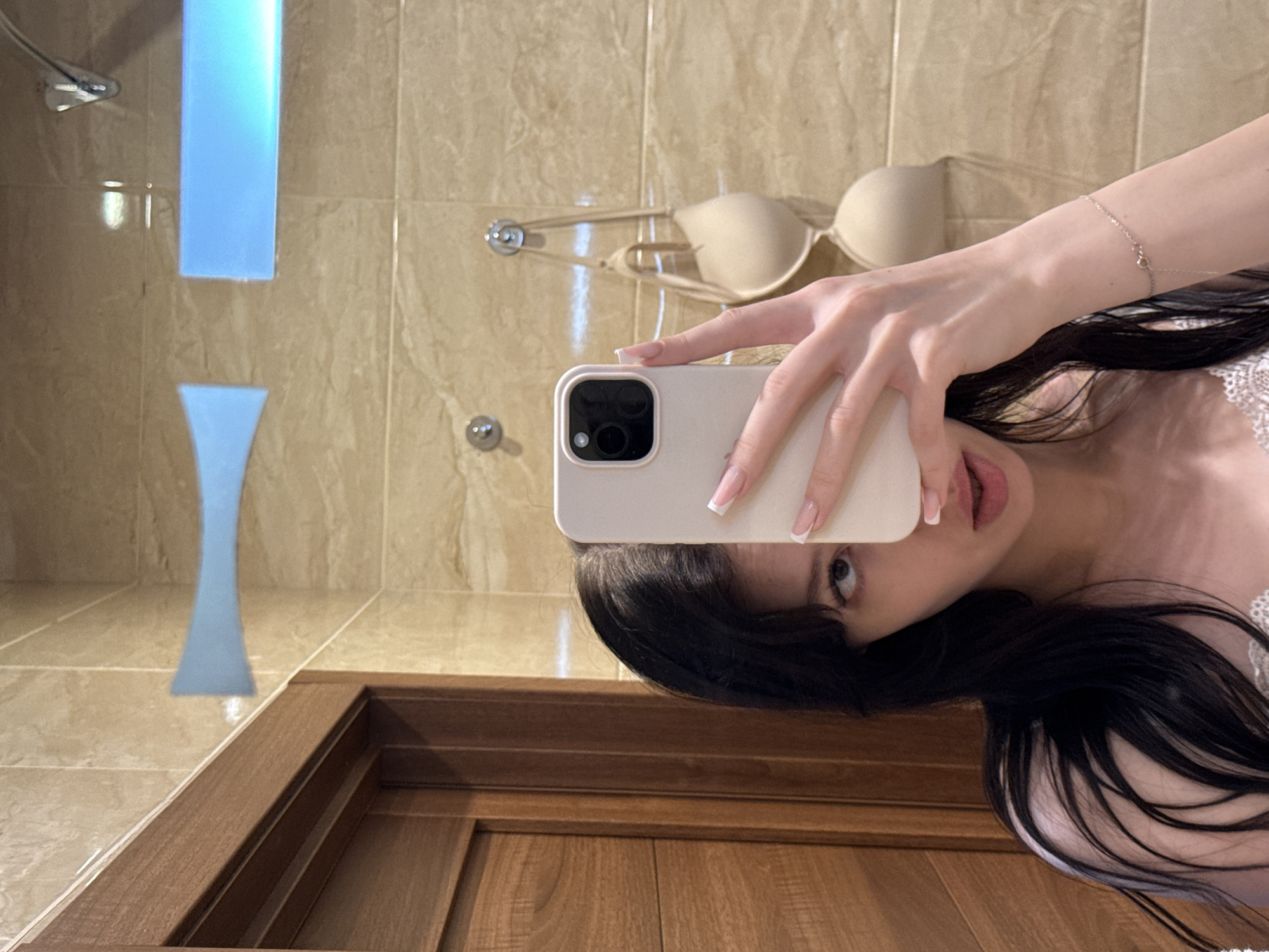 A young woman with long, dark hair takes a selfie in a bathroom. She is holding a smartphone in her right hand, obscuring part of her face. She is wearing a white lacy top. In the background, a beige bra is hanging from a hook on a tiled wall. A wooden door is visible on the left side of the image. There are also two blue light fixtures on the tiled wall above the bra. The overall impression is a private, domestic setting.