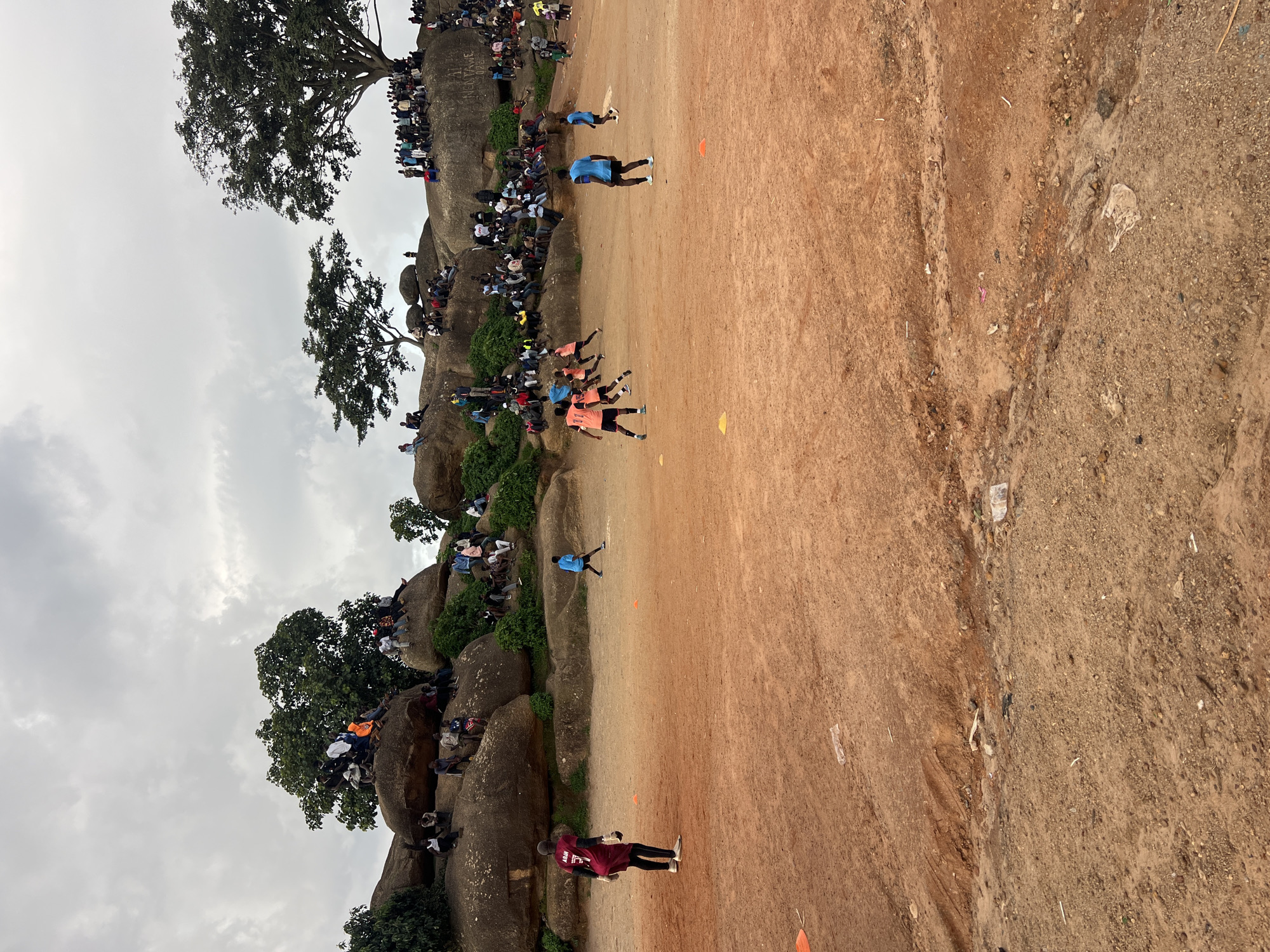 The image captures a lively scene in Jos, Nigeria, likely during the daytime under a cloudy sky. A group of people are gathered on and around large rock formations, some of which are covered in greenery. These individuals appear to be spectators, observing an event unfolding on a dusty, reddish-brown field below.
The central activity appears to be a soccer game or a similar sport. Several people are dressed in athletic attire, including jerseys and shorts, and are actively engaged on the field. One person in a blue shirt is standing on the field, while others in orange and blue jerseys are in motion, suggesting they are playing. A few individuals in maroon and black are also visible on the field.
The spectators are perched on the rocks, forming a natural amphitheater. Their diverse clothing suggests a casual gathering. The overall atmosphere seems engaged and possibly excited, with people leaning forward and watching the game intently. The presence of orange cones on the field indicates a demarcated playing area. The terrain is rough, with visible rocks and sparse vegetation on the slopes, characteristic of an outdoor, possibly rural or semi-urban, location. There is no visible text in the image.