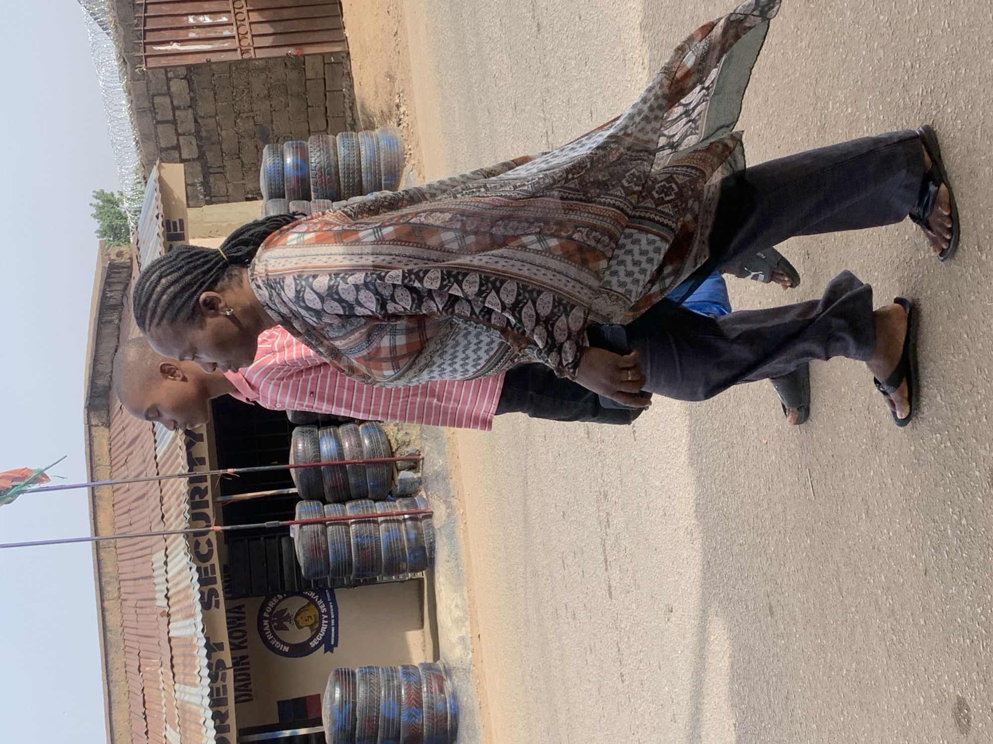 Two people are standing in front of the "Nigerian Forest Service Dadin Kowa Security" building. The building is on the left side of the image, with tires stacked in front of it. One person is wearing a patterned top and dark pants with sandals. The other person is wearing a striped shirt and dark pants. The location is Jos, Nigeria, based on the sign on the building.