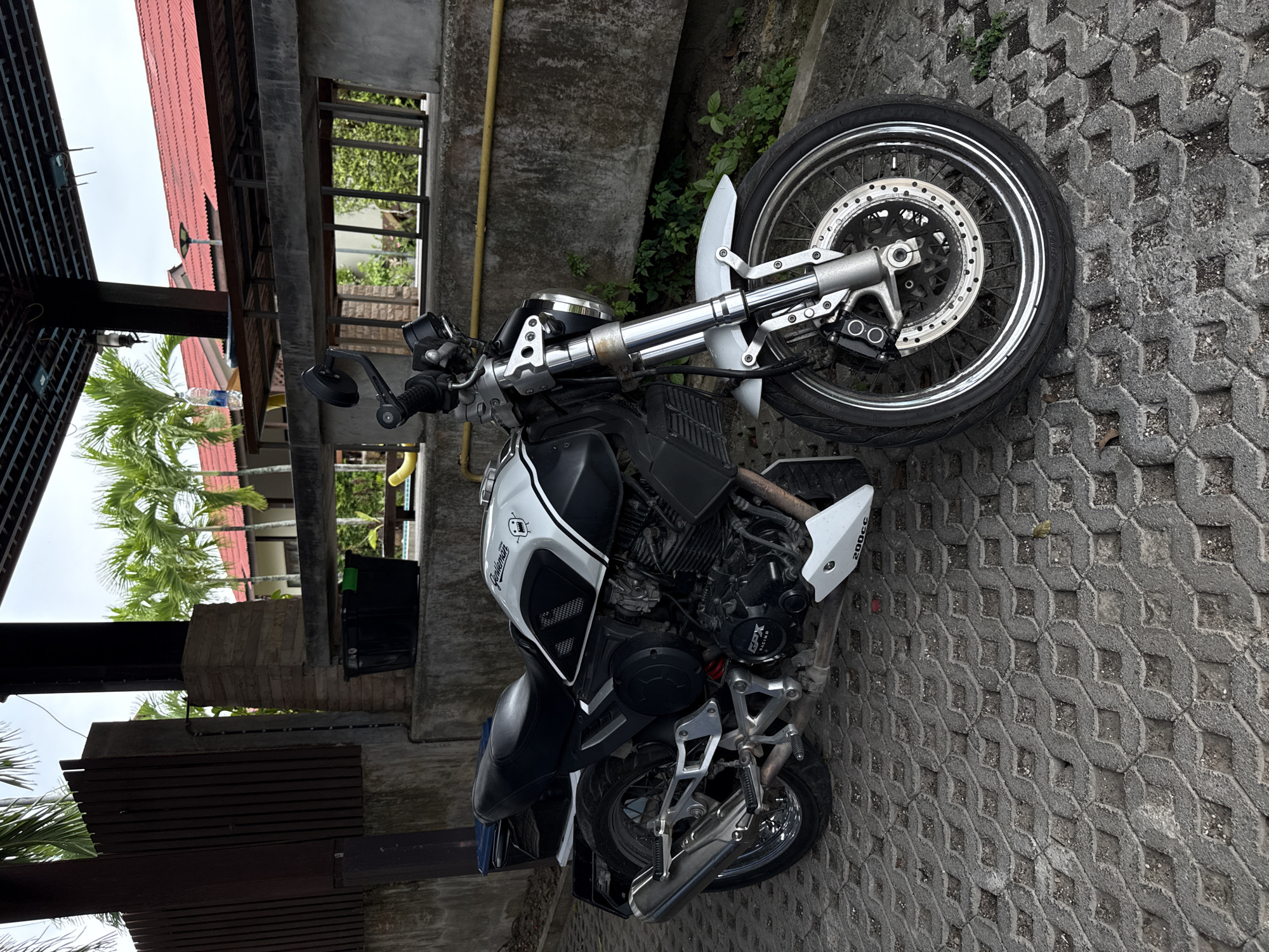 An outdoor, eye-level, full shot shows a parked motorcycle in Baan Bo Phut, Thailand, on a gray, patterned pavement. The motorcycle is black and white with silver accents and has a small white fender. It is positioned to the left of the frame, angled toward the right. Behind the motorcycle, a rough concrete wall runs horizontally across the scene. Above the wall, a covered structure with a dark roof and exposed beams is visible. Several palm trees with green fronds are seen behind the structure, and red-tiled roofs of buildings are in the background. The sky is overcast and light gray.