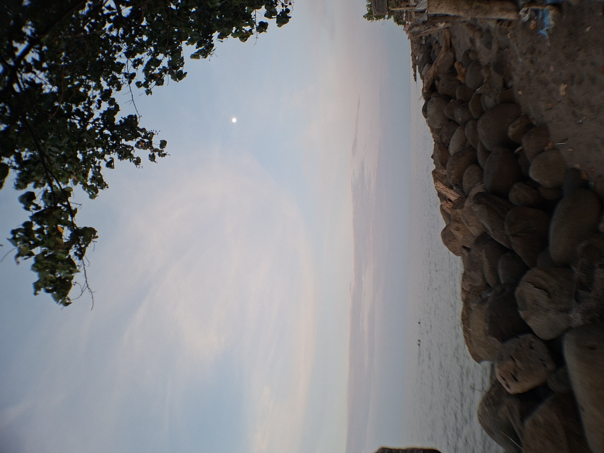 The image shows a view of the sky and ocean from a shoreline in Lhokseumawe, Indonesia. The photo is rotated 90 degrees clockwise. On the left, leaves and branches from a tree obscure a portion of the sky, which transitions from blue to a pale pink hue near the horizon. A small, bright celestial body, likely the moon, is visible in the upper-left of the sky. The ocean is visible along the right side, bordered by a rocky shoreline made of large, rounded stones. Several small objects, potentially birds or boats, are visible on the ocean surface. On the top edge are some man-made structures.