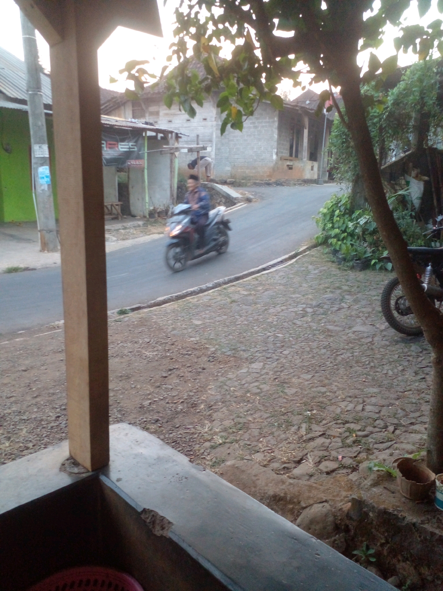 The image depicts a rural scene in Pungangan, Indonesia, likely taken during the late afternoon or early evening given the lighting. In the foreground, a wooden post and part of a concrete structure suggest a porch or covered area. The ground is covered in gravel and irregular paving stones. A person is riding a dark-colored motorcycle on a paved road that curves to the right. The motorcycle has its headlights on. The road is bordered by a curb and a drainage ditch. Beyond the road, there are several buildings with brick and tiled roofs, typical of residential structures in the area. Green foliage and trees are visible, with one tree's branches and leaves prominently in the upper right portion of the frame. Another motorcycle is parked on the right side of the frame, partially obscured by a tree. A small woven basket and a portion of a bucket are also visible near the parked motorcycle.