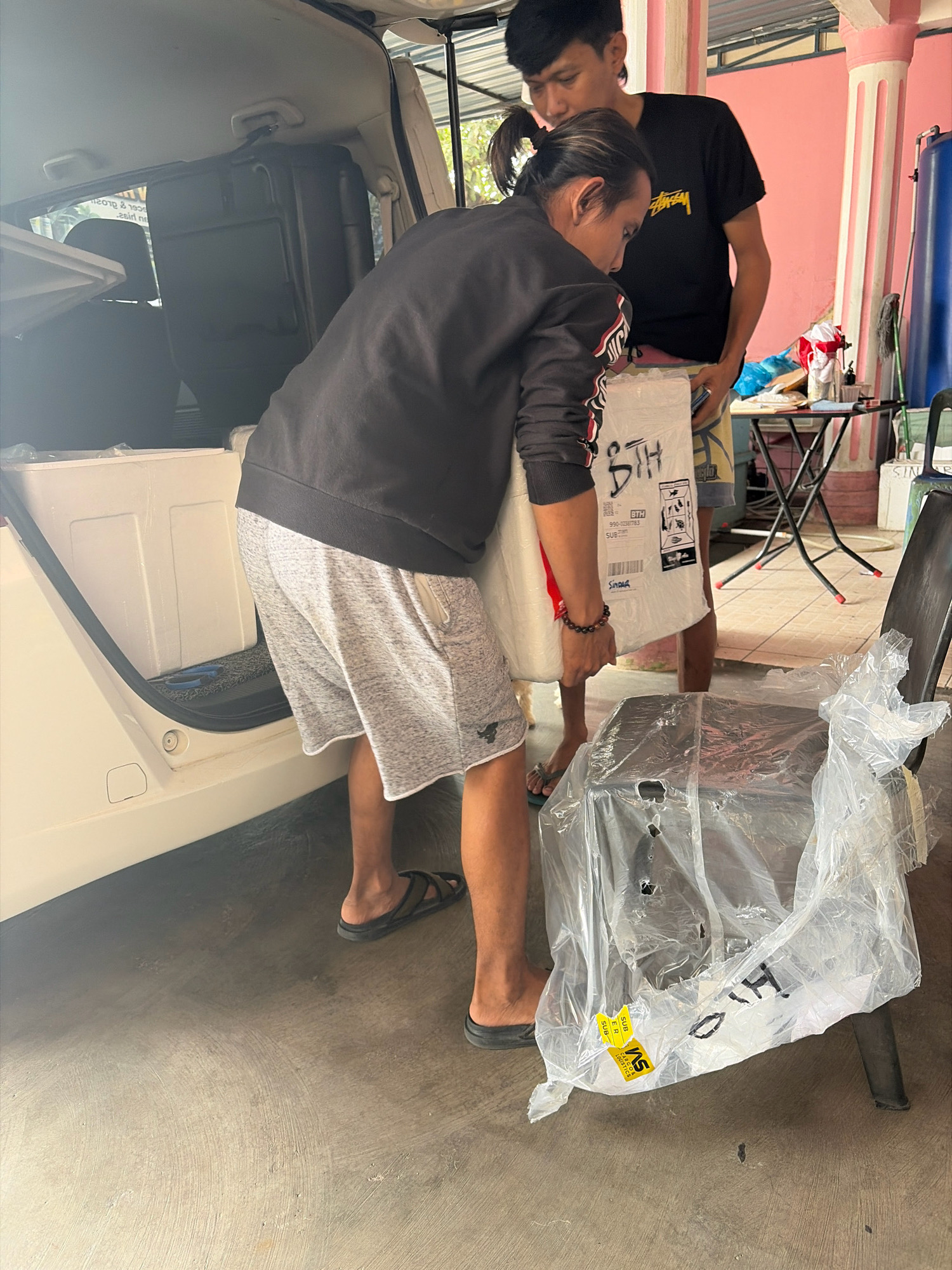 Two individuals are engaged in moving a large, white, rectangular package from the open rear of a white car. One person, wearing a black jacket and gray shorts, is positioned on the left and is partially obscured by the package, holding it from the side. The other person, wearing a black t-shirt and light-colored shorts, stands to the right, assisting with the package. In the foreground, a black chair is covered in clear plastic sheeting. The scene appears to be outdoors, with a concrete floor and a structure with pink and white pillars in the background. To the right, a small table with items on it is visible. The environment suggests a residential or commercial area in a tropical climate.