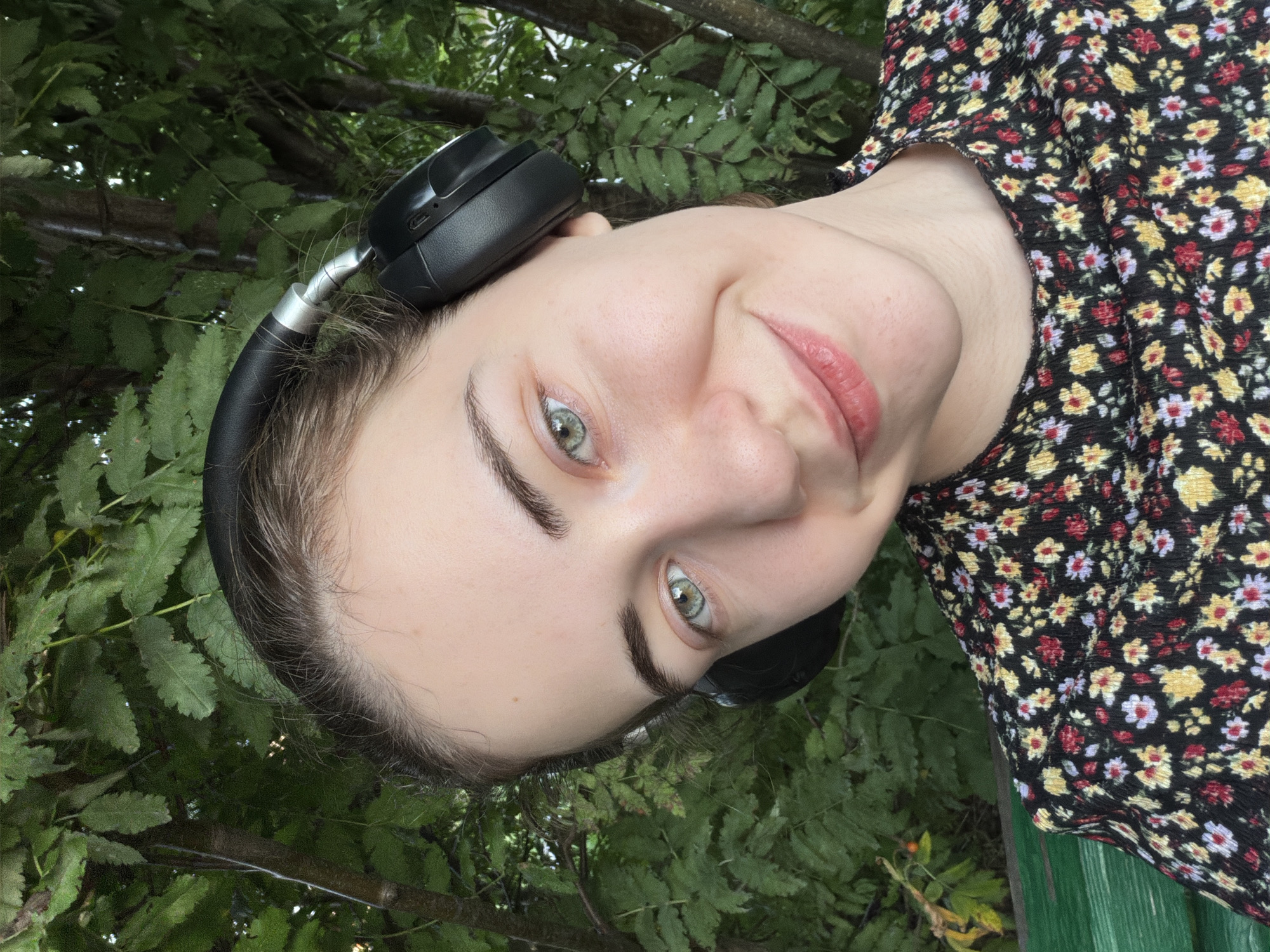 A young woman with light skin, brown hair, and green eyes is pictured from the chest up. She is wearing black over-ear headphones and a black floral-print shirt. Her head is tilted to the right, and she is smiling. The background is a dense foliage of green leaves, suggesting an outdoor setting, possibly a park or garden. There is a hint of a green bench in the lower left corner. The overall lighting suggests it is daytime. The image does not contain enough information to determine the specific location within Zavodoukovsk, Russia.