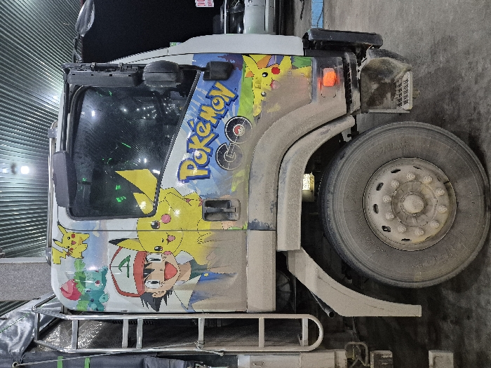 The scene is located in South Hoai Nhon Ward, Vietnam. The image displays the driver's side profile of a white truck cab, heavily soiled with light brown dirt and dust on its lower half. A large, dirty tire with a multi-lug hub is prominent in the lower center. The cab features a prominent, multicolored graphic decal depicting Ash Ketchum, multiple instances of Pikachu, and Bulbasaur, along with the "Pokémon GO" logo. A metal ladder structure, partially obscured by a dark tarp, is visible behind the cab's left side. The cab's side window reflects internal elements and the external environment. An amber side marker light is illuminated on the truck's lower front corner. The environment appears to be an indoor or covered space, characterized by a corrugated metal ceiling with visible linear light sources and a dark, possibly concrete, floor. No individuals are present in the static scene.