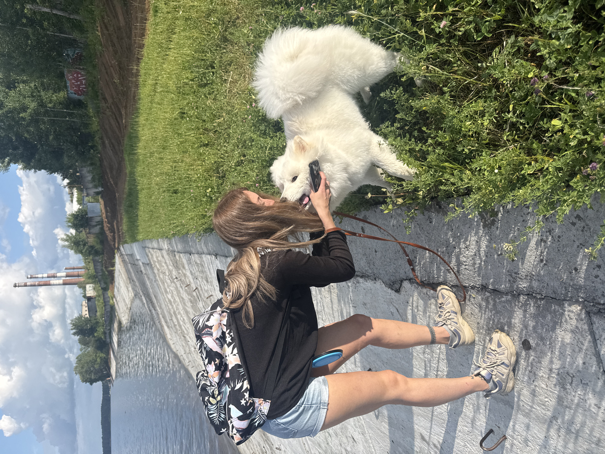 A young woman with blonde hair wearing denim shorts and a black long-sleeved shirt is interacting with a white Samoyed dog on a sunny day. She is holding a mobile phone up to the dog's face, and the dog appears to be licking the phone. The woman is leaning down, and her backpack with a floral pattern is visible on her back. They are standing on a concrete embankment next to a body of water. In the background, there are industrial chimneys, trees, and a grassy slope with some graffiti on a wall. The location is near the Kama River in Chaikovsky, Russia.