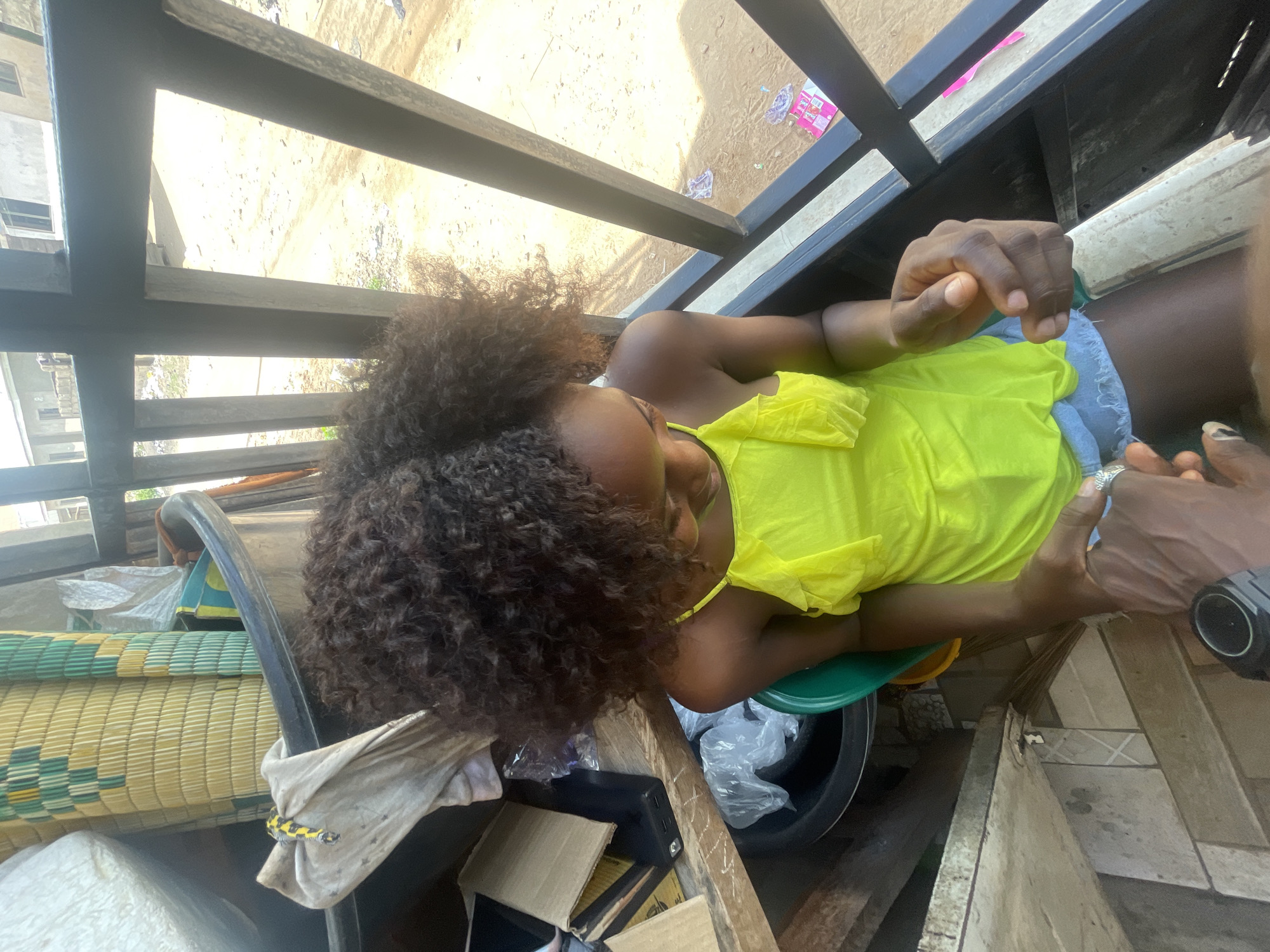 The image depicts a woman with curly hair wearing a yellow top and light blue jeans seated on a green stool. A man's hands are visible, with a smartwatch on his wrist. The setting seems to be on a porch or balcony, as indicated by the black metal railing. Below the railing, the ground appears sandy, and there's visible litter. The surrounding area includes a building facade and mats stacked on the left. In the immediate vicinity of the woman are a black bucket, plastic bags, and discarded cardboard boxes. The location is Lokoja, Nigeria.