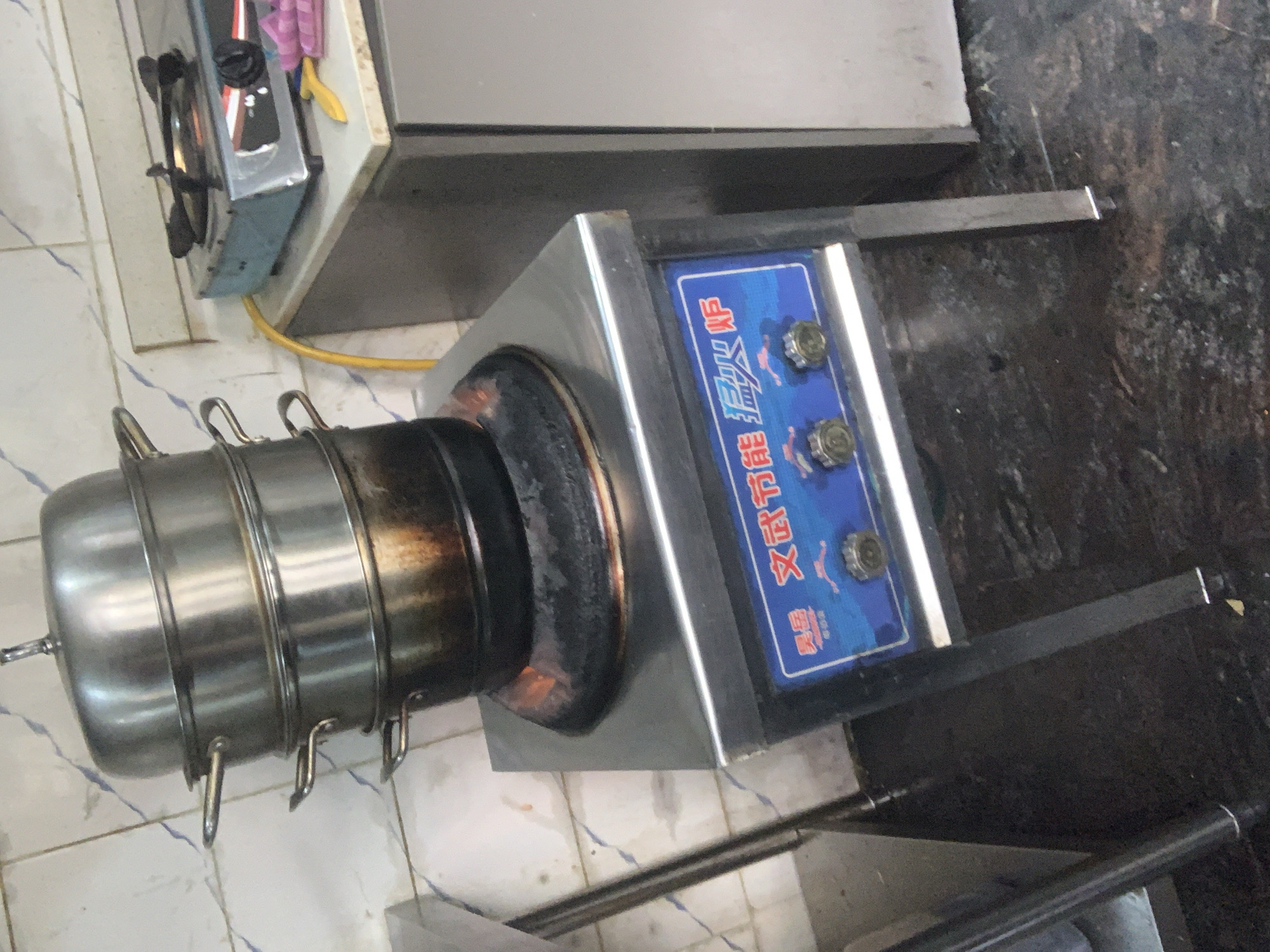 A stainless steel steaming pot is placed on top of a gas burner, which is lit with a visible orange flame, indicating it is in use. The pot appears to be a multi-tiered steamer. The burner is part of a stainless steel stovetop that is integrated into a larger stainless steel appliance with a blue control panel featuring Chinese characters and three control knobs. The appliance is situated on the floor, supported by four legs, and appears to be in a commercial kitchen environment, as suggested by the tiled wall in the background and the general industrial look of the equipment. To the right of the stovetop, a portion of a stainless steel cabinet is visible, and on top of it, a small blue gas stove with a flame and a yellow gas hose is present. Also on top of the cabinet are some pink and yellow objects, possibly kitchen utensils. The floor is dark and appears to be made of concrete or a similar industrial material, with some debris or stains visible. The overall scene depicts a functional kitchen setup, likely in a restaurant or food establishment, in Abuja, Nigeria, where cooking activities are actively taking place.