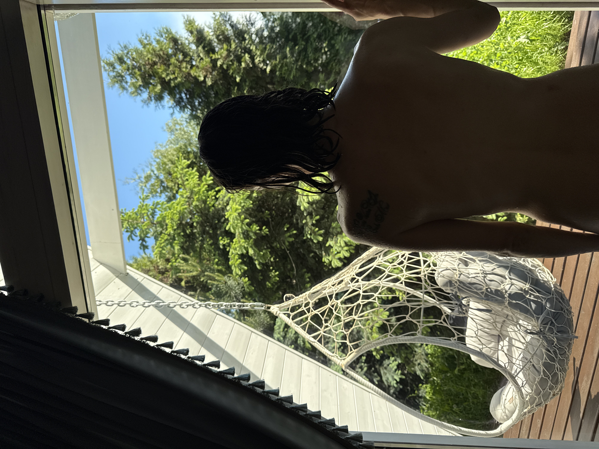 A person with dark, wet hair stands inside, near a window overlooking an outdoor scene in Чёрное, Russia. The person's back is visible, revealing a tattoo on their left shoulder. Outside, there are trees and a blue sky. A hanging chair with white cushions is suspended within the frame, and part of a window blind is visible in the lower left corner. The lighting suggests it is daytime.