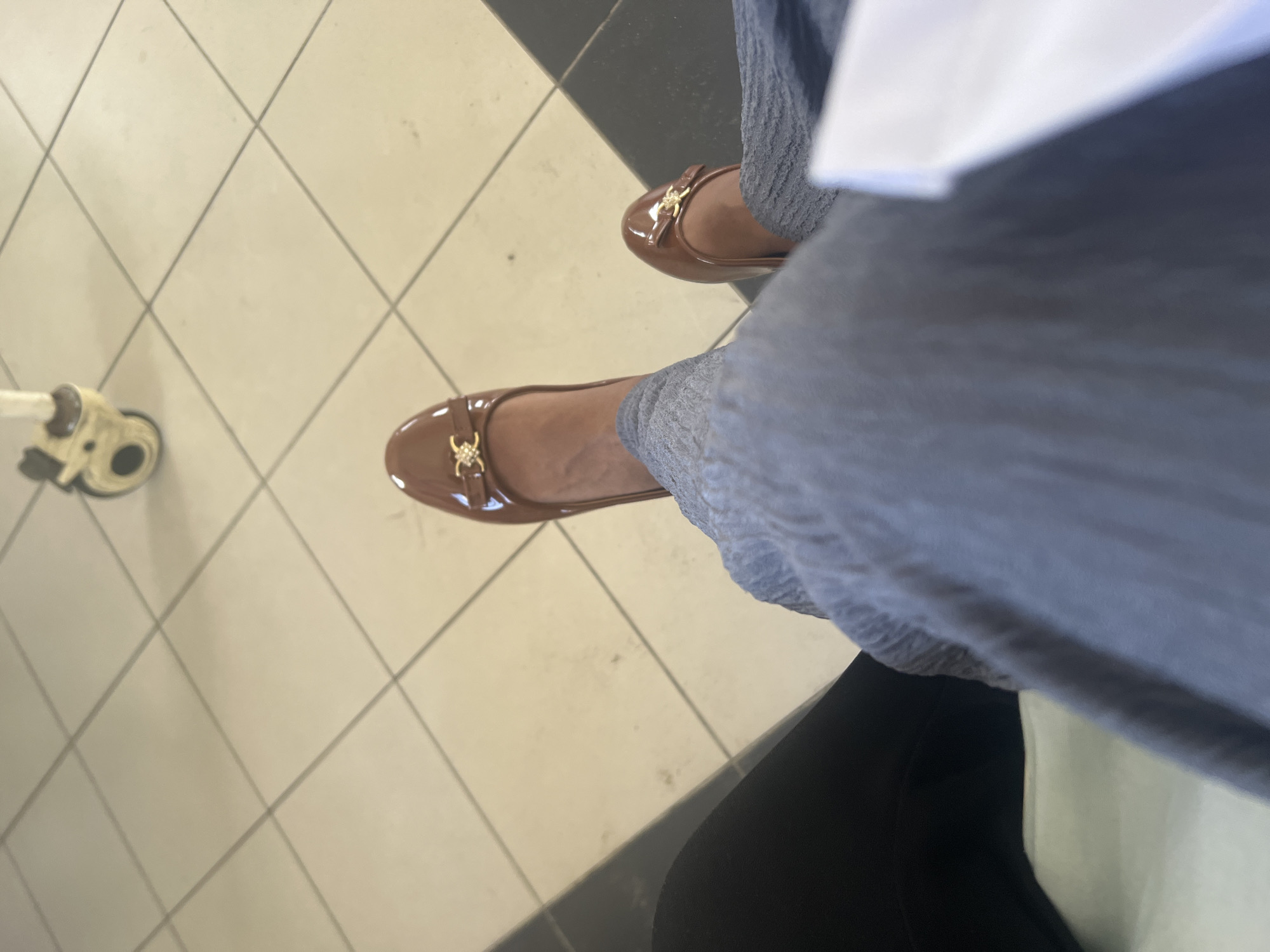A person is wearing brown ballet flats with gold embellishments and gray pants, sitting on a chair. The floor is covered in white and black tiles. There is a small white medical device with a nozzle is in the lower left of the image. The location is most likely in Tanzania, as requested. Given the presence of the medical device, the location could be a clinic or hospital waiting area in Dar es Salaam, Tanzania.