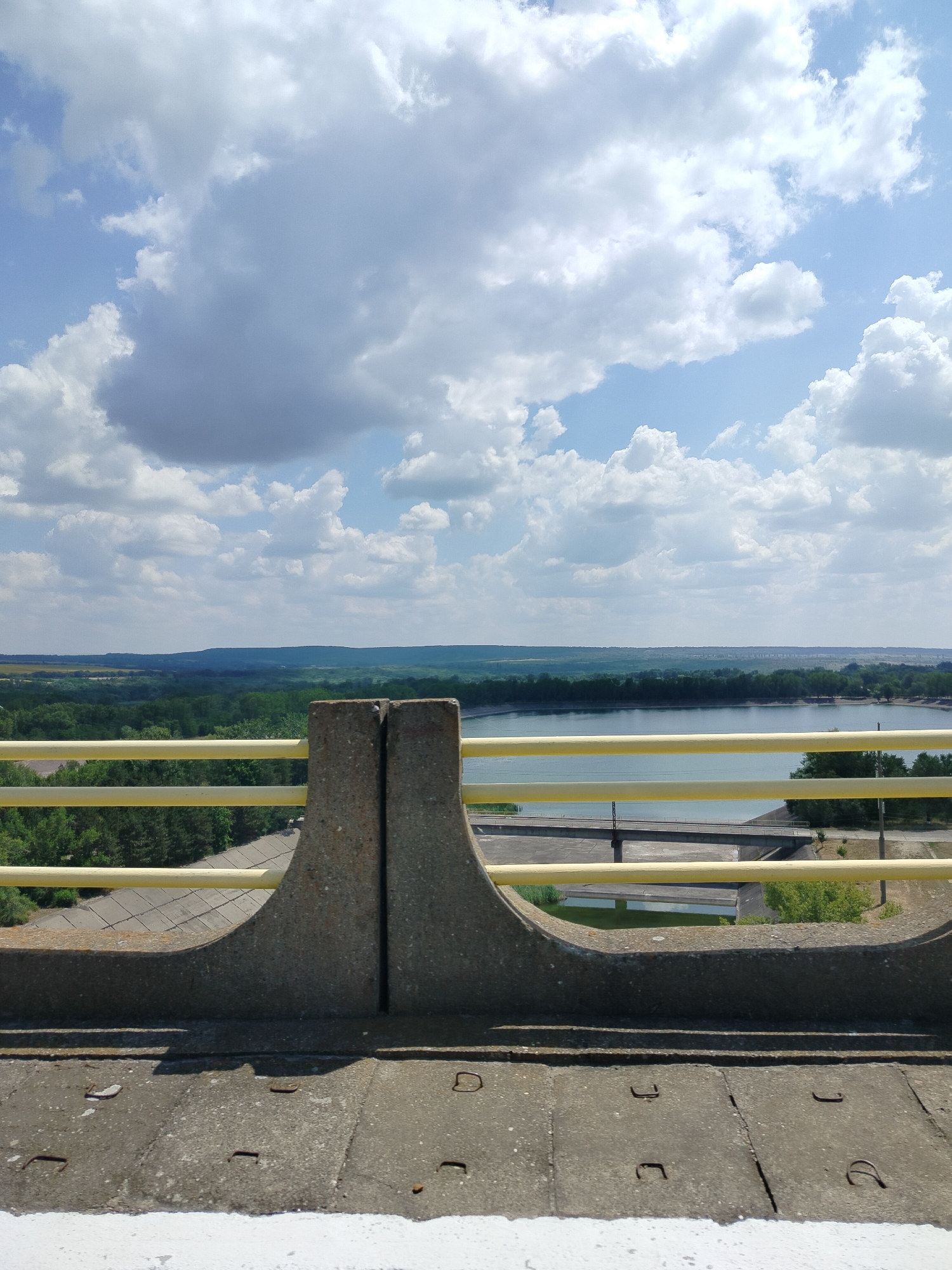 The image depicts a scenic view of the Ștefănești reservoir in Romania, captured from a vantage point likely atop a dam or bridge. The foreground is dominated by a concrete structure, part of a railing, with two distinct concrete pillars displaying a vertical fissure between them. Two horizontal yellow bars, typical of a safety railing, extend across the frame, separating the viewer from the expanse beyond.
The mid-ground reveals the reservoir, its surface a calm, light blue, reflecting the sky. Beyond the reservoir, a lush green landscape unfolds, characterized by dense forests that transition into rolling hills in the distance. A bridge or roadway is visible crossing the water in the mid-ground.
The sky above is a vibrant blue, adorned with a dramatic display of fluffy white and grey clouds, suggesting a partly cloudy day. The overall impression is one of natural beauty, with the man-made structures subtly integrated into the environment. The image appears to be a still photograph, with no people or active events depicted.