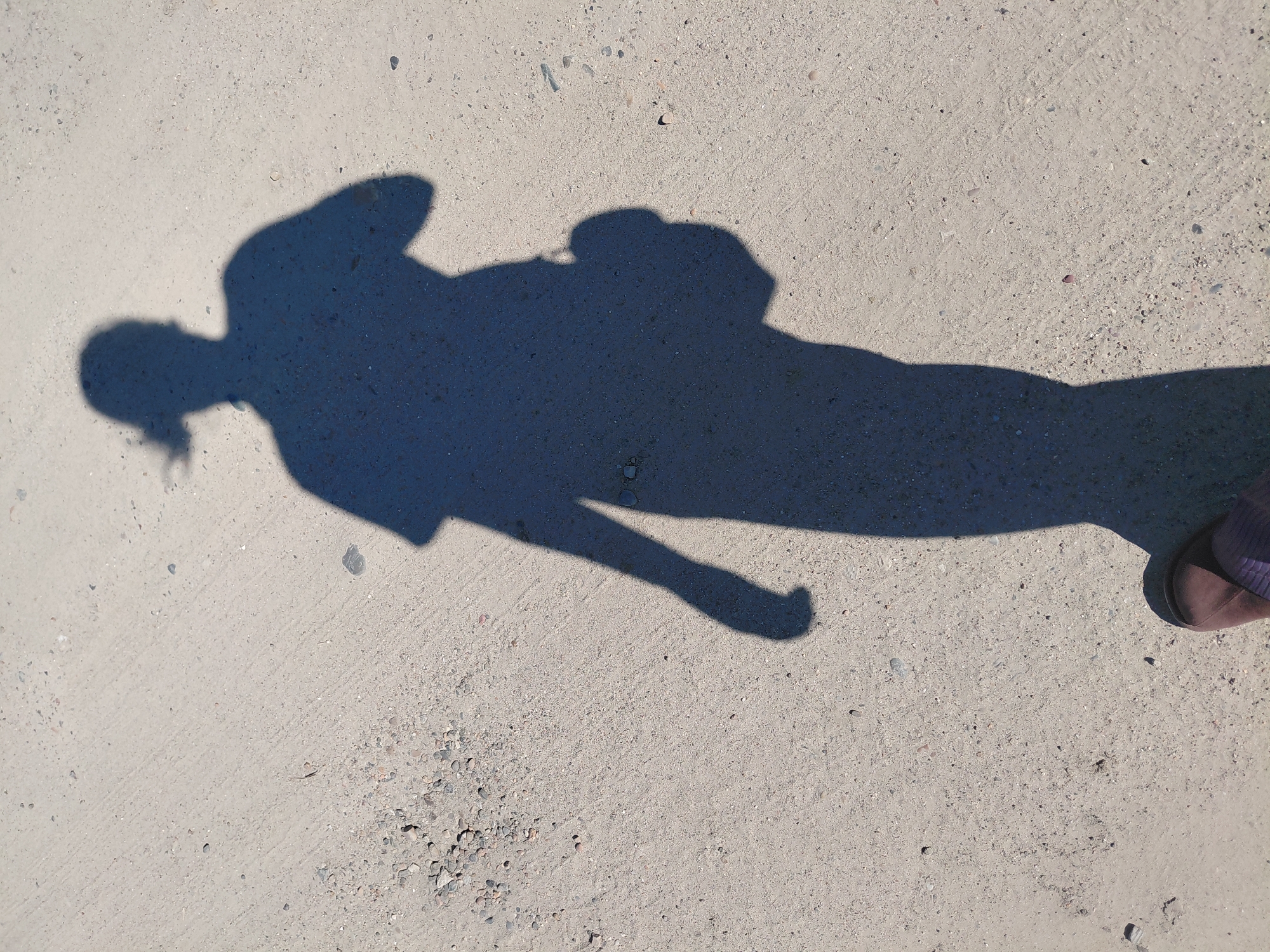 The image shows a shadow of a person walking on a dusty, grey, outdoor surface. The surface appears to be a path or road with small stones and gravel. The shadow indicates the person is wearing a t-shirt and has their hair tied back. They are also carrying a bag over one shoulder. The shadow is cast by a light source from above and to the right. The bottom right corner of the image shows a glimpse of the person's foot wearing a dark purple sandal. The image is taken from a slightly elevated angle, looking down at the ground and the shadow.