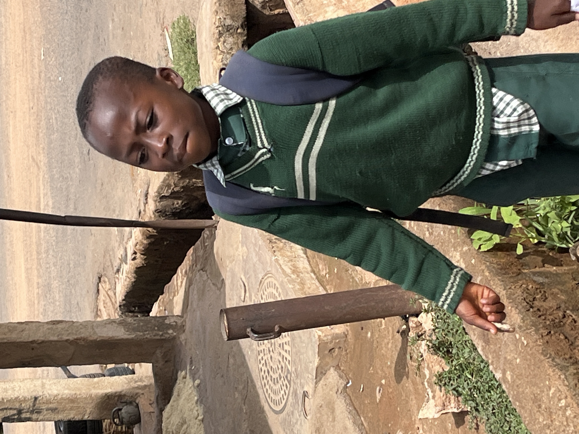 A young, dark-skinned boy is standing on a concrete structure, potentially a culvert or drainage system, near a paved road. He is wearing a dark green sweater with white stripes and a collared shirt underneath, along with dark pants and a dark blue backpack. He is holding a small, light-colored object in his right hand, possibly a piece of stone or a twig. The location appears to be in Jos, Nigeria, based on the likely context and visual cues of the environment. Vegetation grows near the concrete structure. A brown metal pipe is visible near the boy, attached to the concrete. The image is angled slightly, giving a downward perspective.