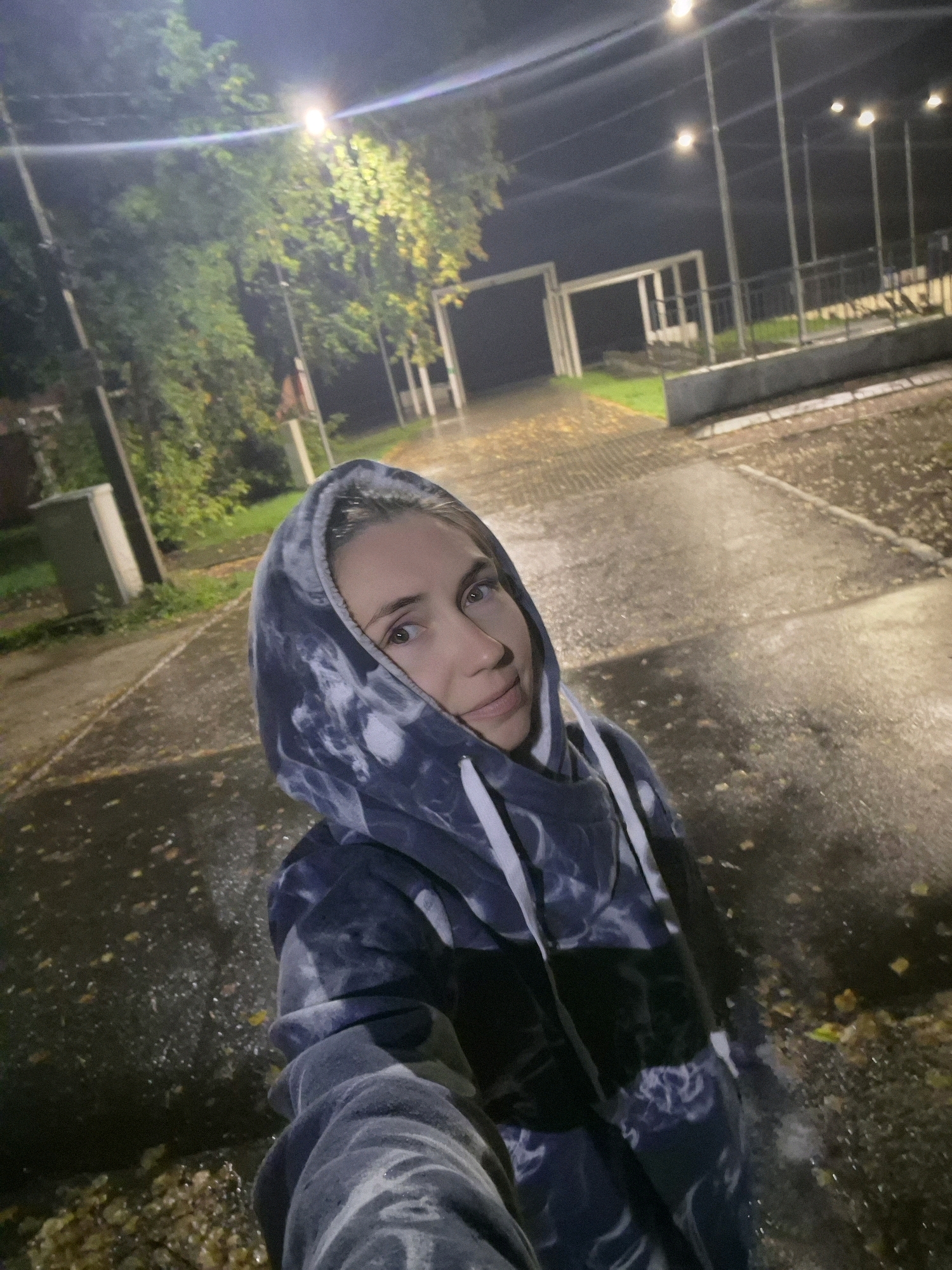 A selfie is taken by a woman outdoors at night, in the city of Chaikovsky, Russia. She is wearing a dark tie-dye hoodie with the hood pulled up. Her face is partially obscured by the hood. The background shows a paved area that appears wet, with fallen leaves scattered on the ground. Streetlights illuminate the scene, casting a glow on trees and structures in the distance. One streetlight in particular is very bright and creates a lens flare in the image. There are also power lines visible against the dark sky. The environment suggests an outdoor public space, possibly a park or a street.