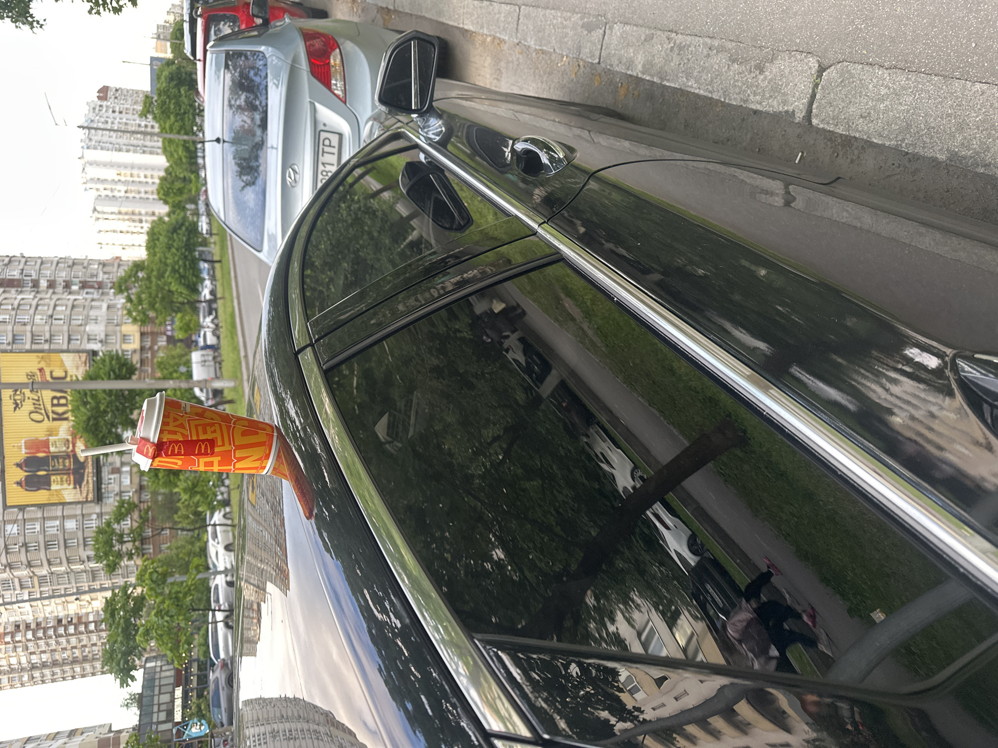 A disposable paper cup with a red lid and straw, emblazoned with orange lettering and the McDonald's logo, rests precariously on the roof of a parked black car. The cup appears to be filled with a beverage. The car is parked on a city street in Kyiv, Ukraine, with other vehicles lined up along the curb. In the background, tall residential buildings dominate the skyline, interspersed with trees and a large billboard advertisement. A person is visible in the reflection of the car's window, walking on the sidewalk. The scene suggests a typical urban environment in Kyiv.