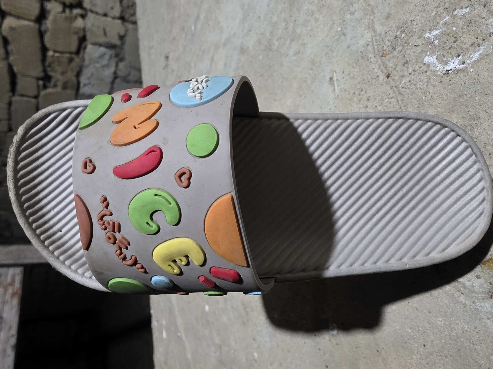 The image shows a close-up, low-angle shot of a single, light gray slide sandal with a patterned sole. The sandal is decorated with various colorful, raised embellishments on its strap. These include brown ovals, green crescent shapes, a yellow curved shape resembling a worm or fungus, a red oval, an orange semi-circle, a pink oblong shape, a light blue circle, and several green circles. Some of these embellishments appear to be cartoonish characters or objects. On a light blue circular emblem on the strap, there are white characters, possibly in a foreign script. The sandal is resting on a concrete surface, with a shadow cast to the left. In the background, a blurred stone wall and a wooden structure are visible. The scene is outdoors, and the lighting suggests it may be nighttime or a shaded area.