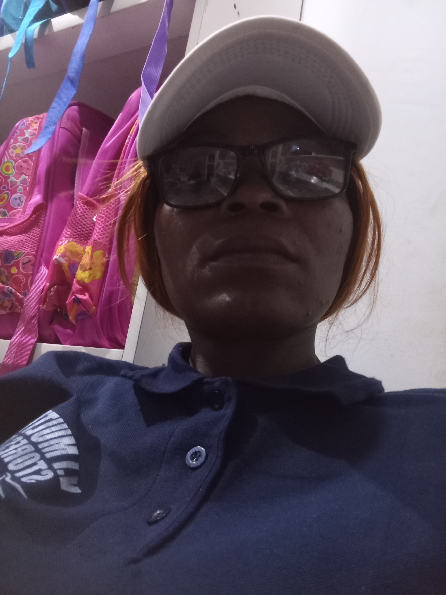 A medium shot, chest-up photograph shows a person from the chest to the top of their head. They are wearing a dark blue polo shirt and a white baseball cap, as well as dark-rimmed glasses. Their hair is visible under the cap and is a reddish-brown color. In the background, pink backpacks and blue straps are hanging, suggesting a setting like a school or storage area. The lighting is dim, and the background is largely out of focus. The scene is indoors, with a plain wall visible on the right side of the frame. The person's expression is neutral.