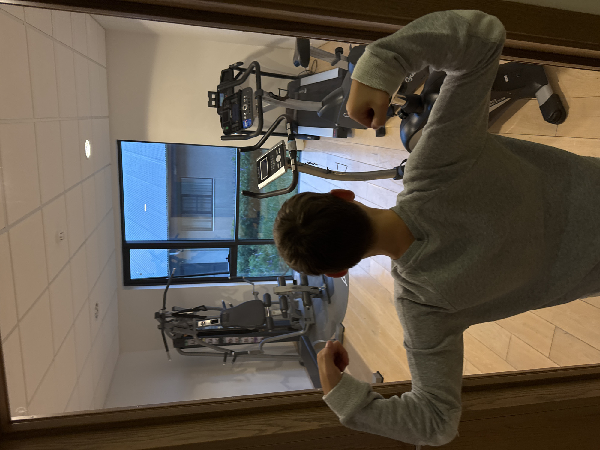 A child wearing a gray sweatshirt is visible in the foreground, partially obscuring a view of a gym through a doorway. The gym contains an exercise bike and a weight machine. Through a window, an exterior view is visible, revealing what appears to be a multi-story building and greenery, suggesting an urban or suburban environment. The scene has no indication to confirm this is Vik, Iceland.