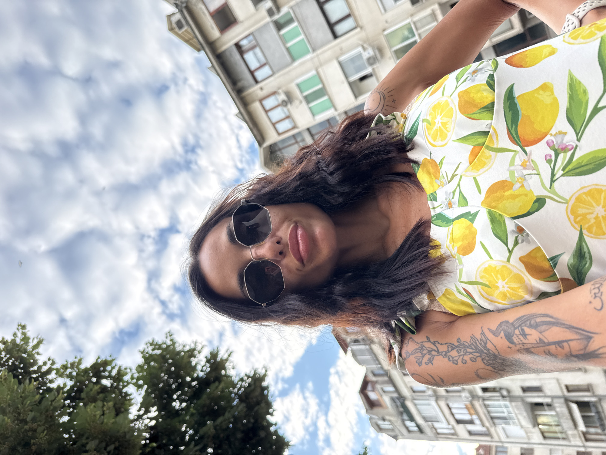 A woman is captured in a selfie taken outdoors in the city of Pleven, Bulgaria. She is wearing sunglasses and a dress with a lemon print. In the background, buildings and a tree are visible under a cloudy sky.