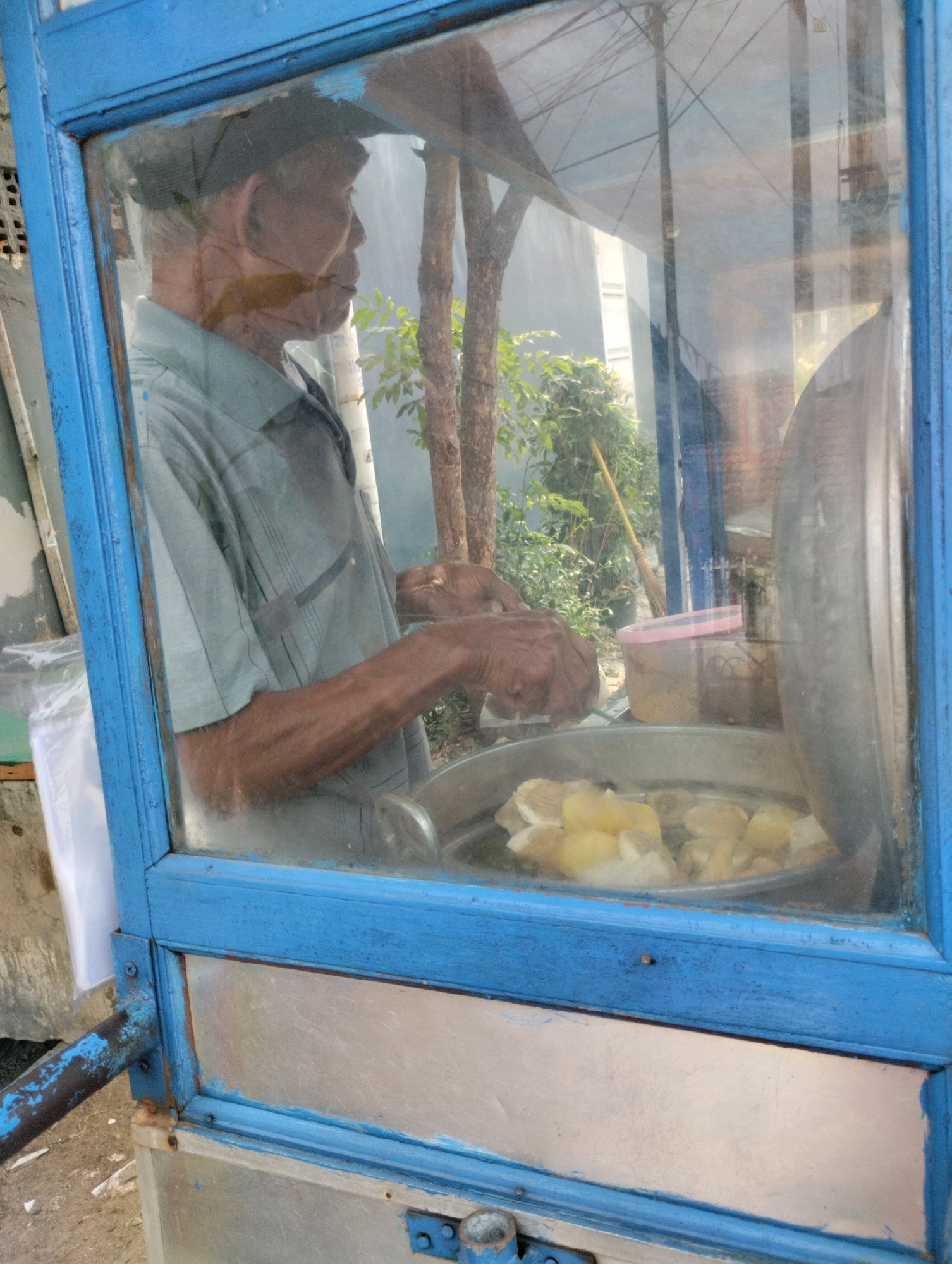 The image depicts an elderly Indonesian man in profile, wearing a light blue collared shirt and a dark cap. He is positioned inside a blue-painted wooden food cart, which has a glass front. His hands are engaged in preparing food within the cart. In front of him, a metal steamer pot contains pieces of what appear to be boiled potatoes. To his right, a pink-lidded plastic container is visible. The background shows some greenery and trees, suggesting an outdoor setting, possibly a street or roadside in Bekasi, Indonesia. The cart appears worn, with peeling blue paint on its frame.