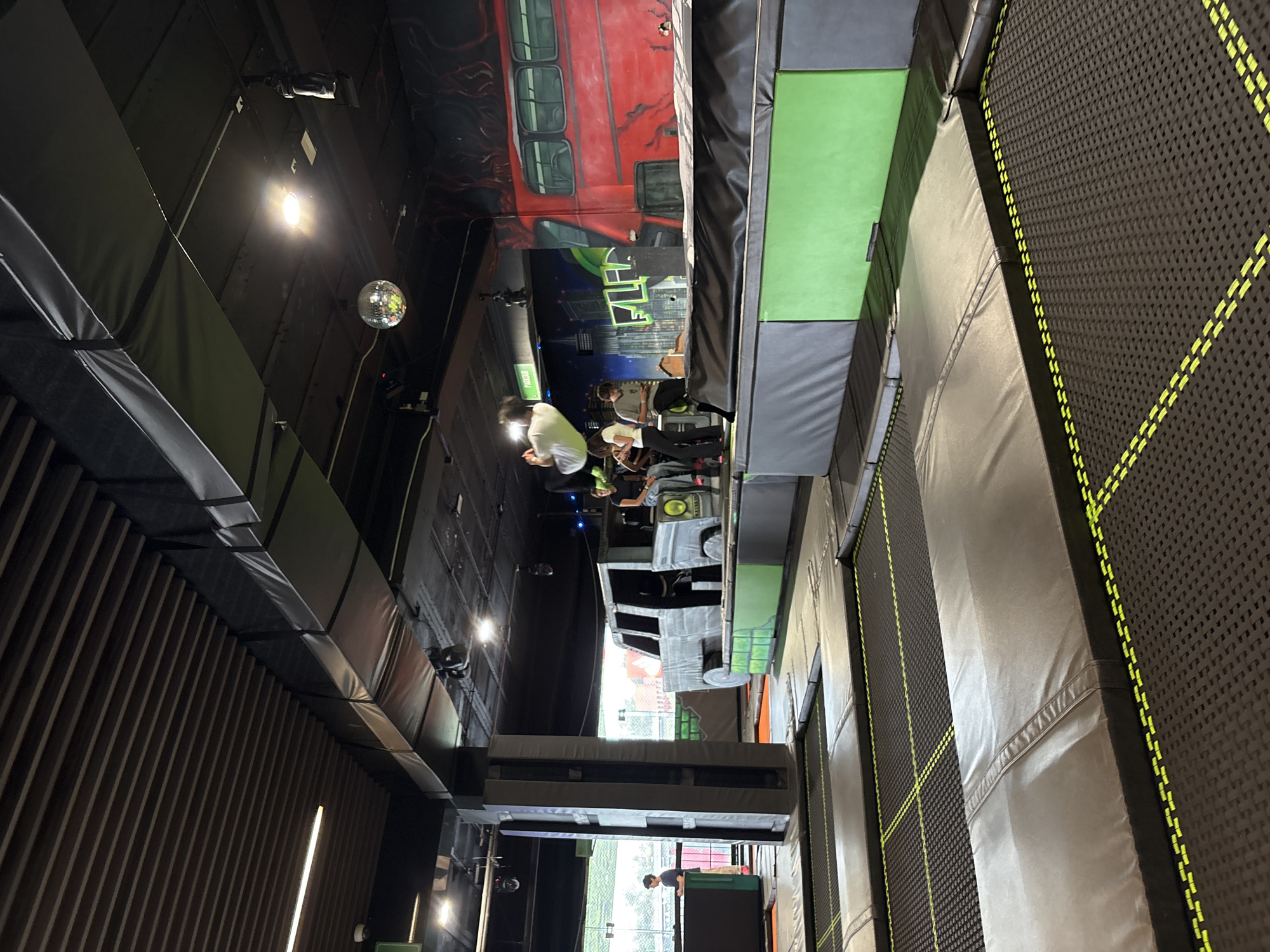 The image shows the interior of a trampoline park in Mexico City, Mexico. The scene includes trampolines with black and yellow mesh and padded edges, wall murals depicting urban scenes and a red double-decker bus, and several people. Some individuals are standing or sitting near the murals, while others are positioned closer to or on the trampolines. Overhead, there is a disco ball and visible lighting fixtures. The walls are lined with dark, vertically-oriented slats and exposed ductwork. Through a window in the background, a glimpse of the outside environment is visible.