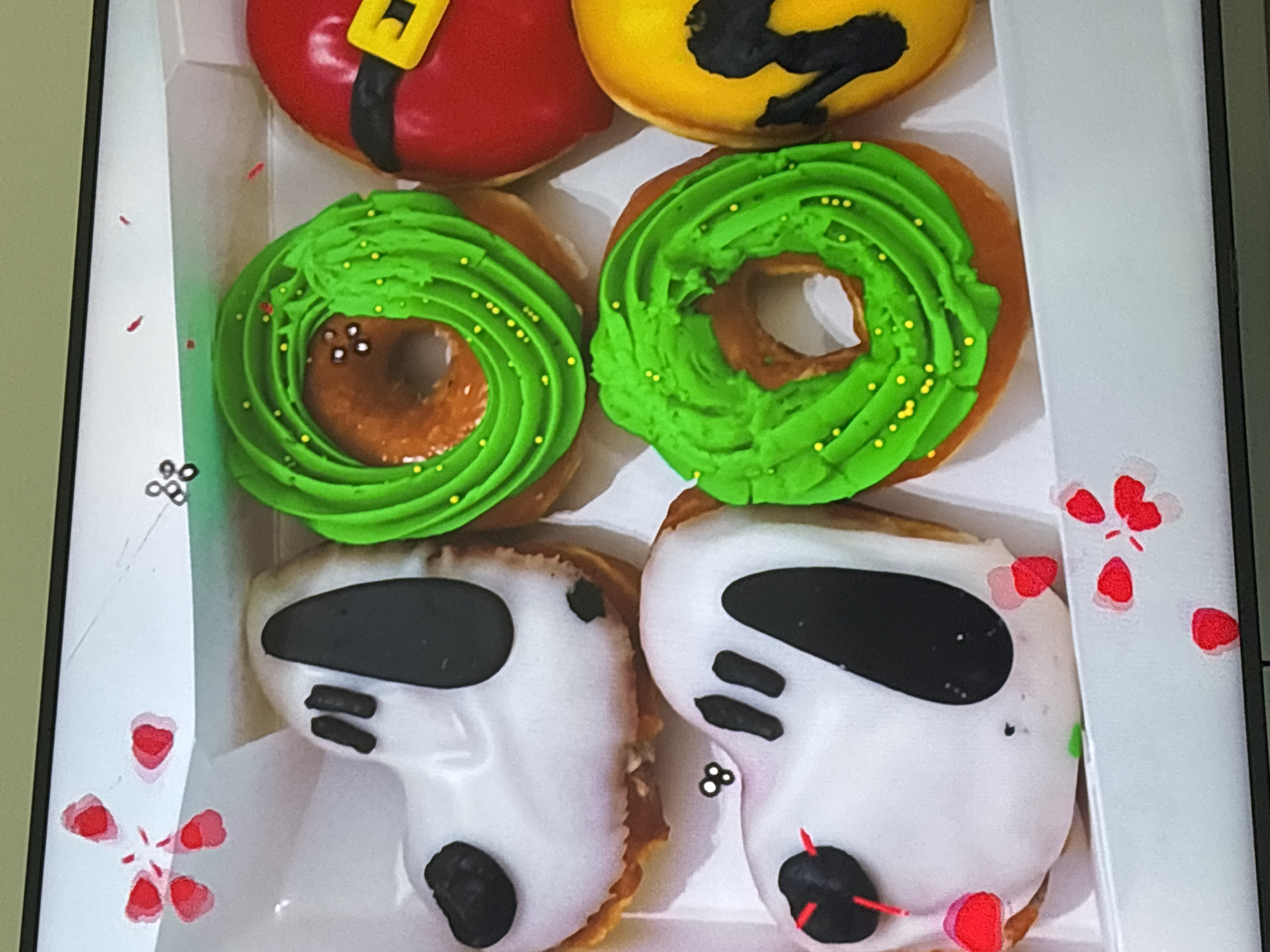 The image shows a close-up of a white cardboard box containing six holiday-themed donuts. The donuts are arranged in two rows of three. In the top left is a donut decorated to resemble Snoopy's head, with white icing, black ears, and black eyes. To its right is a ring donut covered in bright green frosting and small yellow sprinkles, resembling a Christmas wreath. In the top right is a donut decorated as Santa Claus's suit, with red icing, a black belt, and a yellow buckle. In the bottom left is another Snoopy donut, identical to the one above. To its right is another donut resembling a Christmas wreath, identical to the one above. In the bottom right is a yellow donut with black markings, possibly representing a Minion character or another holiday figure. The box appears to be on a flat surface, possibly a table or counter. The background is blurred and out of focus, showing a hint of a television screen with some decorative elements. There are no people or specific Dubai landmarks visible in the image.
