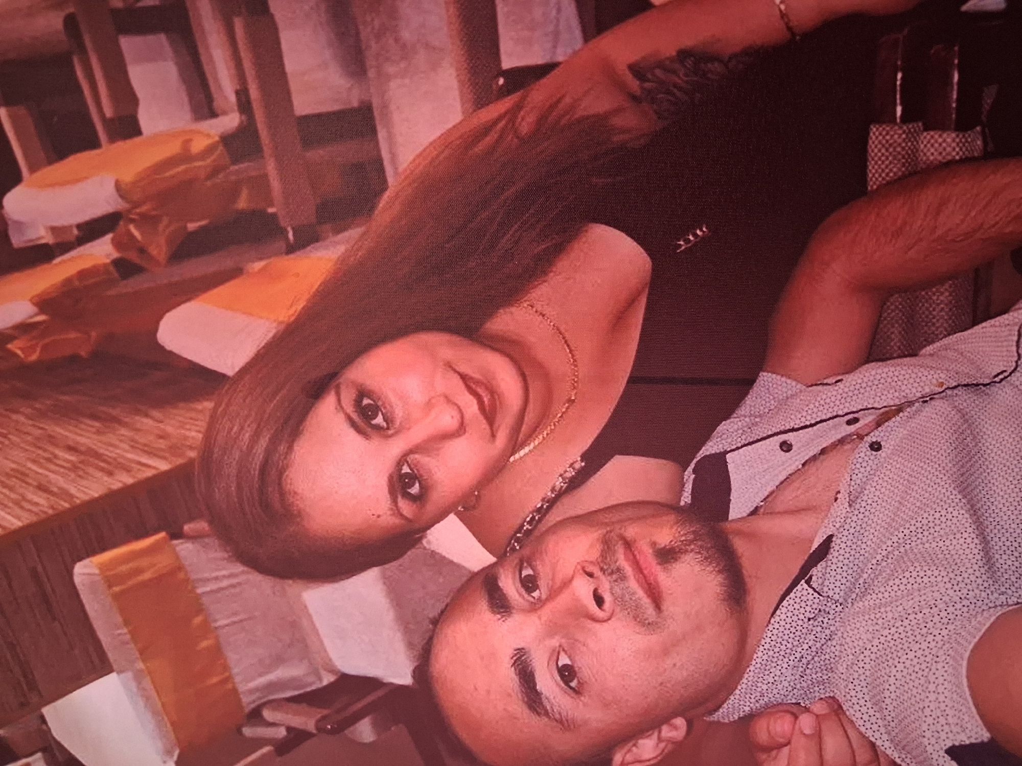 A close-up, slightly blurred, candid outdoor portrait captures a man and a woman posing together. The man, in the foreground, is looking directly at the camera with a faint smile and a goatee. He is wearing a light-colored, short-sleeved collared shirt with a subtle polka dot pattern. The woman is positioned behind and to the right of the man, also looking towards the camera. She has long, dark hair and is wearing a dark, sleeveless top and a delicate gold necklace. Her left arm is resting on the man's shoulder. The background appears to be an outdoor setting with rows of chairs covered in white fabric and adorned with yellow sashes, suggesting a decorated event space or outdoor seating area. The overall lighting is warm, with a reddish hue. The image does not provide any specific information or visual cues that would allow for the identification of the location as Konevets, Bulgaria.