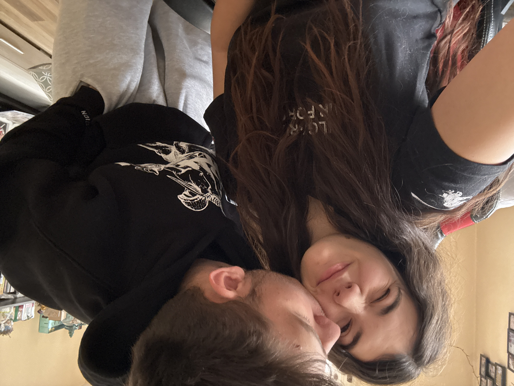 A selfie shows a young adult male and female with their faces close together. The female has long brown hair and wears a black t-shirt with some white letters visible. The male wears a black hooded sweatshirt with a white design on the front. The background is a blurry interior with what appear to be shelves with objects on them. The location is Plovdiv, Bulgaria is not discernible in the photo.