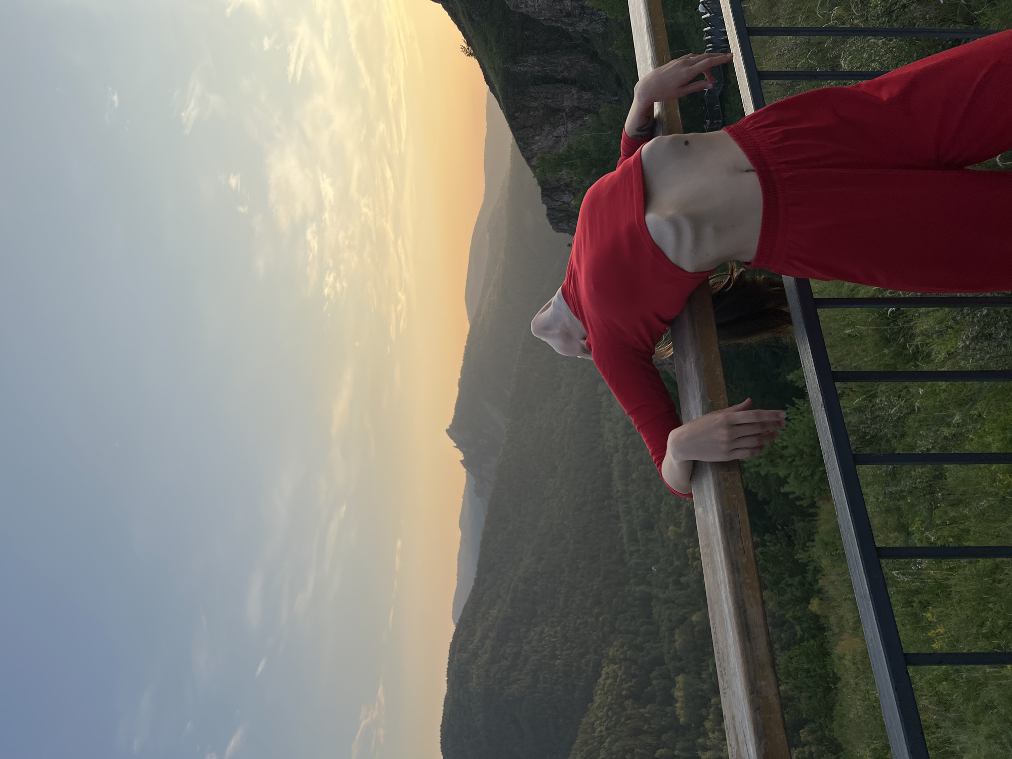 The image captures a young person with a slender build, wearing a red cropped top and red pants, leaning back over a wooden railing. Their head is tilted back, and their body is arched, creating a striking pose against a backdrop of rolling, tree-covered hills and mountains. The setting sun casts a warm, golden glow across the landscape, with soft clouds in the pale blue sky. The foreground features the railing and a dark metal fence, with a hint of green foliage below. This scene is likely taken from an elevated viewpoint, possibly a scenic overlook, within or near the city of Krasnoyarsk, Russia, known for its natural beauty and proximity to the Yenisei River and surrounding taiga.