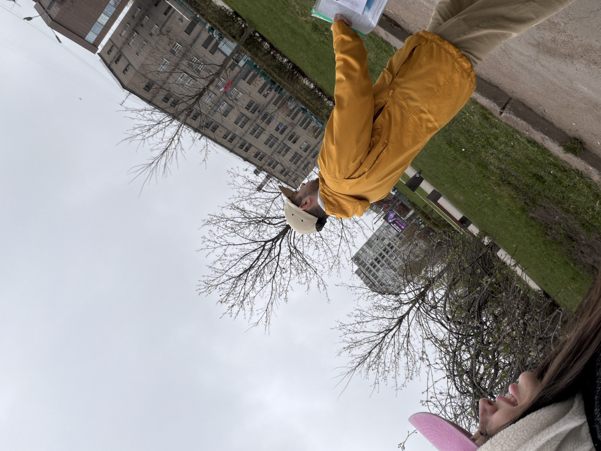 The image, taken in Saint Petersburg, Russia, appears to be a low-angle shot. The sky is overcast. In the foreground, a woman is partially visible, with part of her face and pink hat appearing at the bottom of the frame.
Slightly above her, a man stands on a walkway, wearing a mustard-yellow jacket, khaki pants, and a light-colored baseball cap. He holds a document.
In the background, there are bare tree branches. Behind the trees are gray buildings. Green grass can be seen next to the walkway.