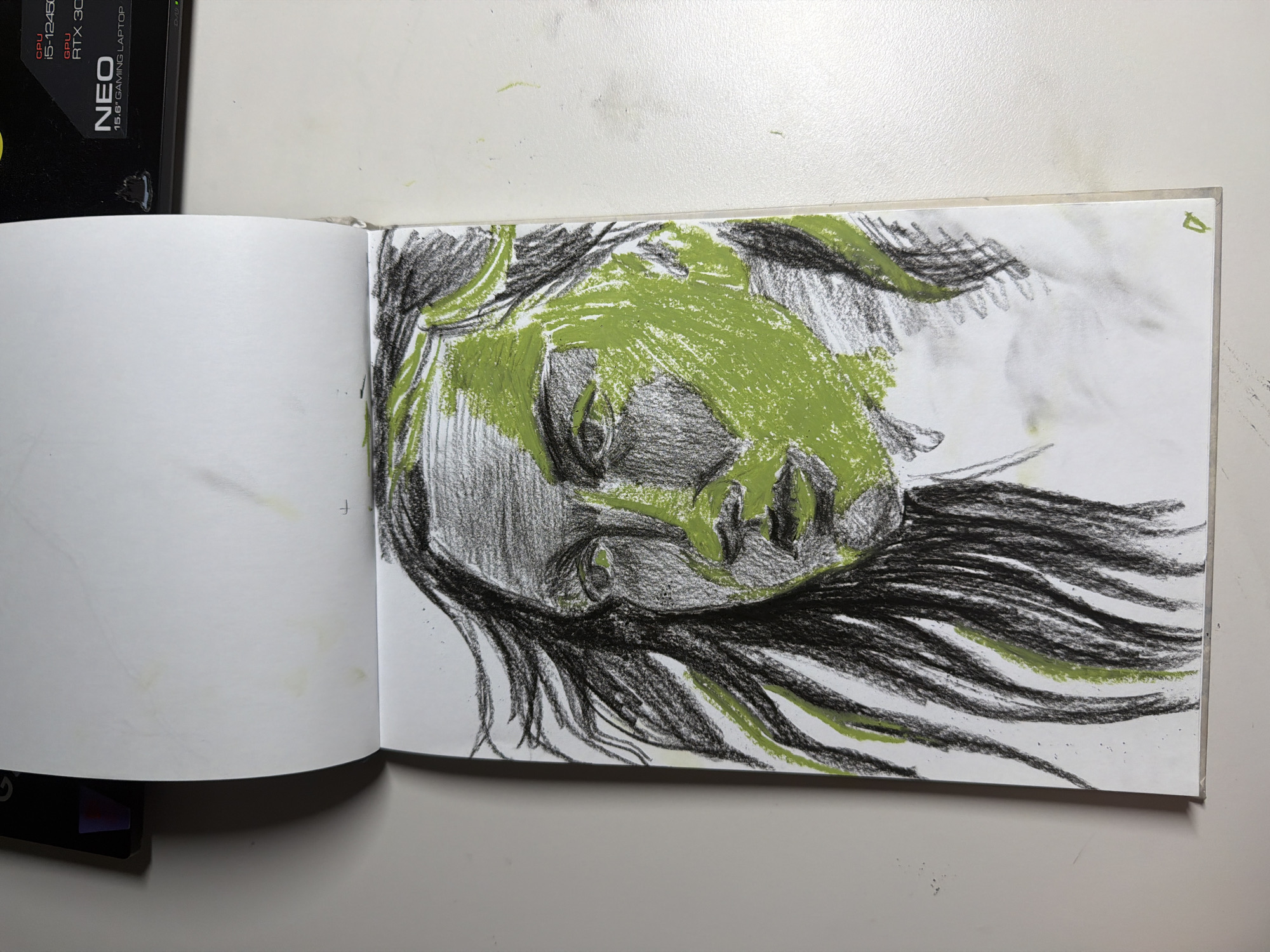 This media file displays a sketchbook open to a page featuring a charcoal and green pastel drawing of a face. The drawing is abstract, with bold, sweeping lines depicting the hair and features of the face, which is rendered in shades of gray and highlighted with vibrant green. The face appears to be angled, with closed eyes and a generally somber or introspective expression. The background of the drawing is mostly white, with some smudges and faint marks.
To the left of the sketchbook, a portion of a black object is visible, bearing text that includes "CPU i5-12450H," "GPU RTX 30," and "NEO 15.6" GAMING LAPTOP." This suggests the sketchbook is placed on or near a gaming laptop.
The scene is indoors, on a white surface, possibly a desk or table, in Dongducheon-si, South Korea. There are no clear indications of the time of day or weather. The focus is on the artwork itself.