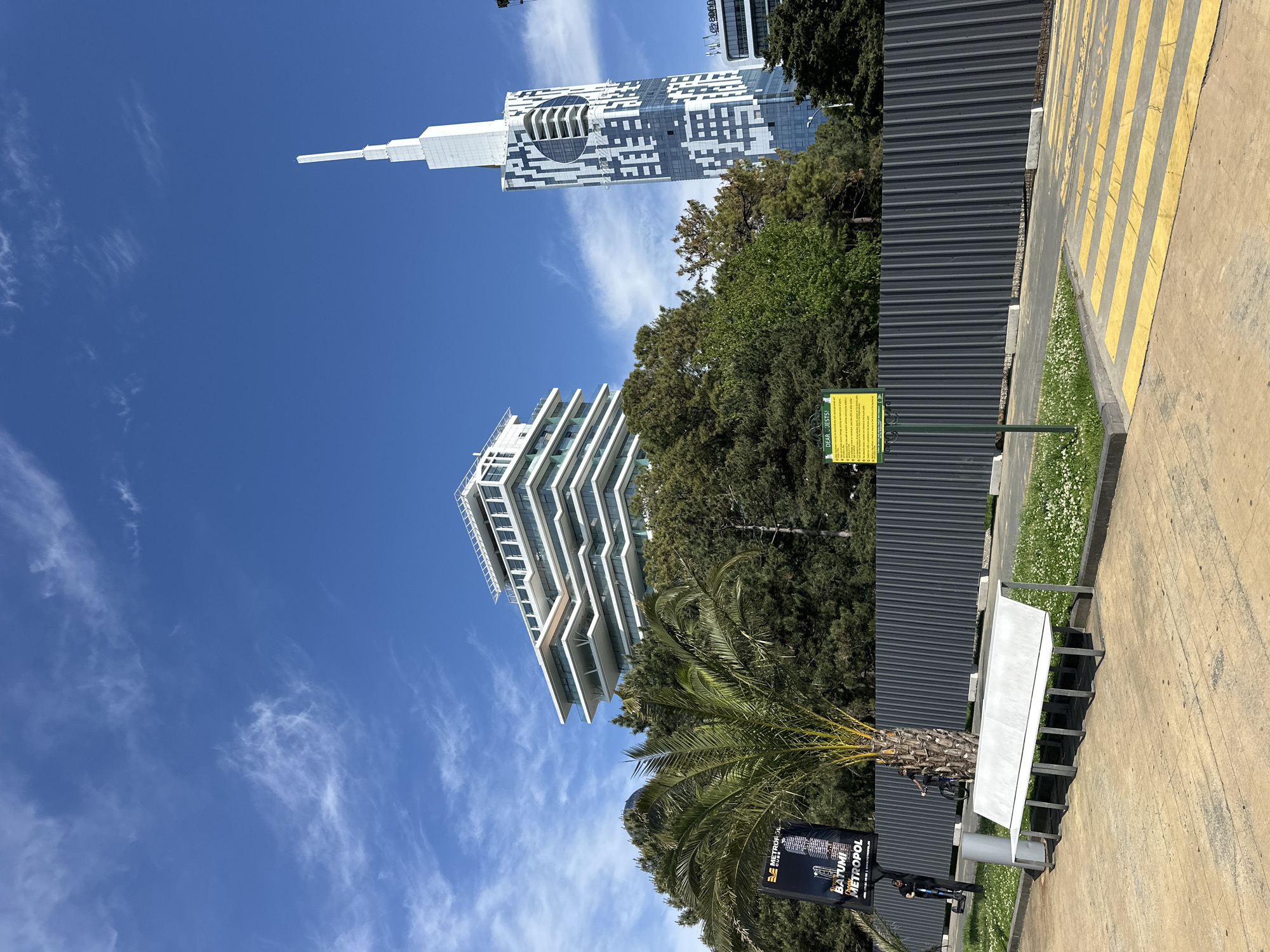 The image captures a sunny daytime view in Batumi, Georgia. The sky is a clear blue with scattered wisps of cirrus clouds. Two modern buildings dominate the background: one is a white, multi-tiered, stepped structure, and the other a taller tower with a geometric black and white pattern and a spire at the top. Green trees fill the space between and around the buildings. In the foreground is a grey corrugated metal fence running parallel to a sidewalk marked with yellow striped lines. A sign post shows an advertisement for a commercial venture called Batumi Metropol. A yellow sign with an unclear message is next to a green bush.