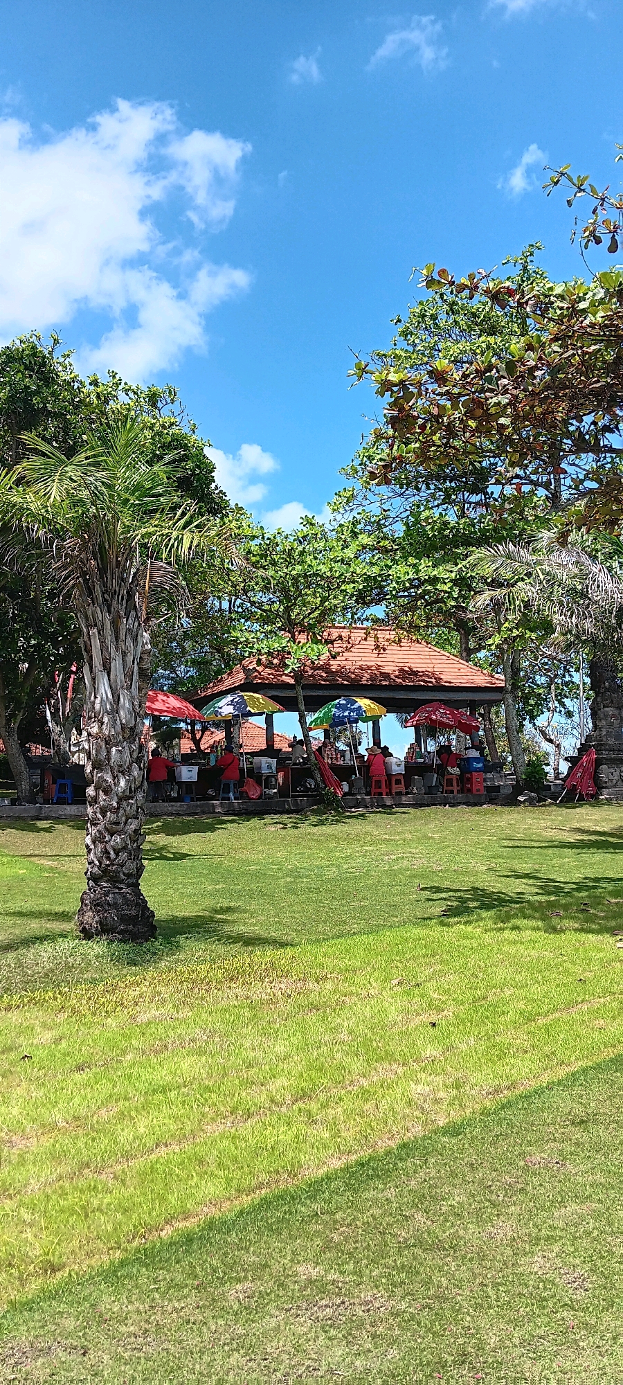 The image depicts a park-like area with a grassy lawn in the foreground. In the middle ground, a shaded pavilion with a terracotta tiled roof is visible, under which several people are gathered. Colorful umbrellas are set up, and there appear to be food or drink stalls. Palm trees and other lush green foliage surround the pavilion. The background shows a clear blue sky with scattered white clouds. The scene suggests a relaxed outdoor setting, possibly a tourist spot or a public recreational area. The overall impression is one of a bright, sunny day in a tropical or sub-tropical environment. The location is stated to be in Kediri, Indonesia.