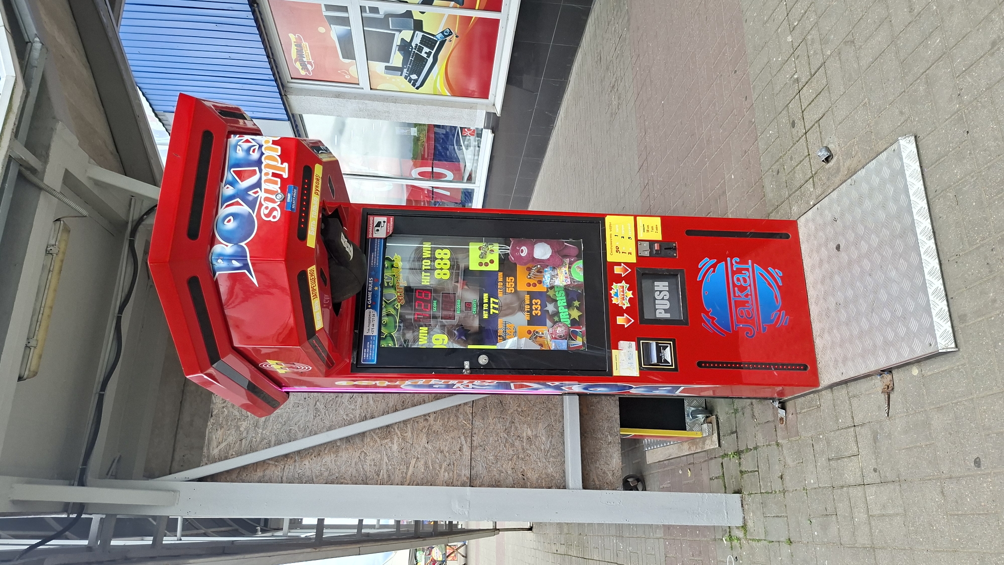 A red boxing arcade game machine, labeled "BOXER SURPRISE," stands on a gray paved sidewalk in front of a building with blue and white panels. The machine has a glass display showcasing prizes and game information, a coin slot, and a large "PUSH" button. To the left of the machine, a ramp with a metal grating leads up to a wooden structure. The overall scene appears to be an outdoor area in the city of Maladzyechna, Belarus.