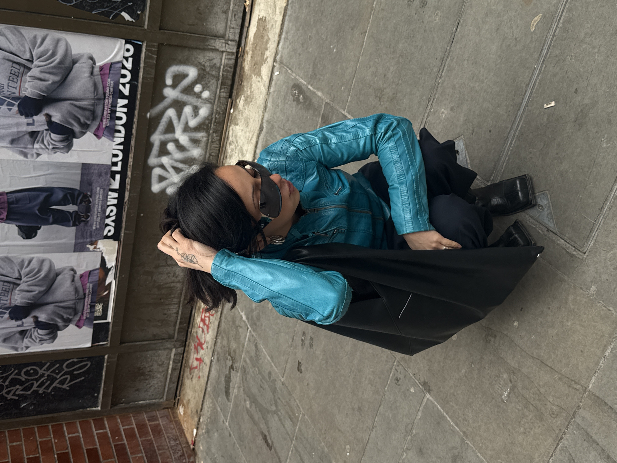 A person is captured from a high angle, sitting on a grimy, paved urban sidewalk, leaning slightly against an unseen surface. They wear a vibrant turquoise blue leather jacket with quilted details on the shoulders and arms, dark trousers, and black square-toed shoes. Large black sunglasses obscure their eyes, and their dark hair frames their face. One hand, adorned with a delicate floral tattoo on the back, rests on their head, while the other holds a large black tote bag that sits on the ground next to them.
The background features a brick wall partially covered by wooden panels, displaying multiple posters. Visible text on the posters includes "SASW LONDON 2020", "www.LIVEFASTOREYOUNG.COM", and "STYLED BY VENEDA CARTER", along with fragments like "NTEED". The wall and ground are marked with white graffiti, with one clear word resembling "2224" and another that appears to be "খাদার" (Bengali for 'food' or 'fodder'). The overall setting suggests a casual moment on a London street, with a mix of street art and commercial advertising. The lighting indicates it is daytime, possibly overcast.