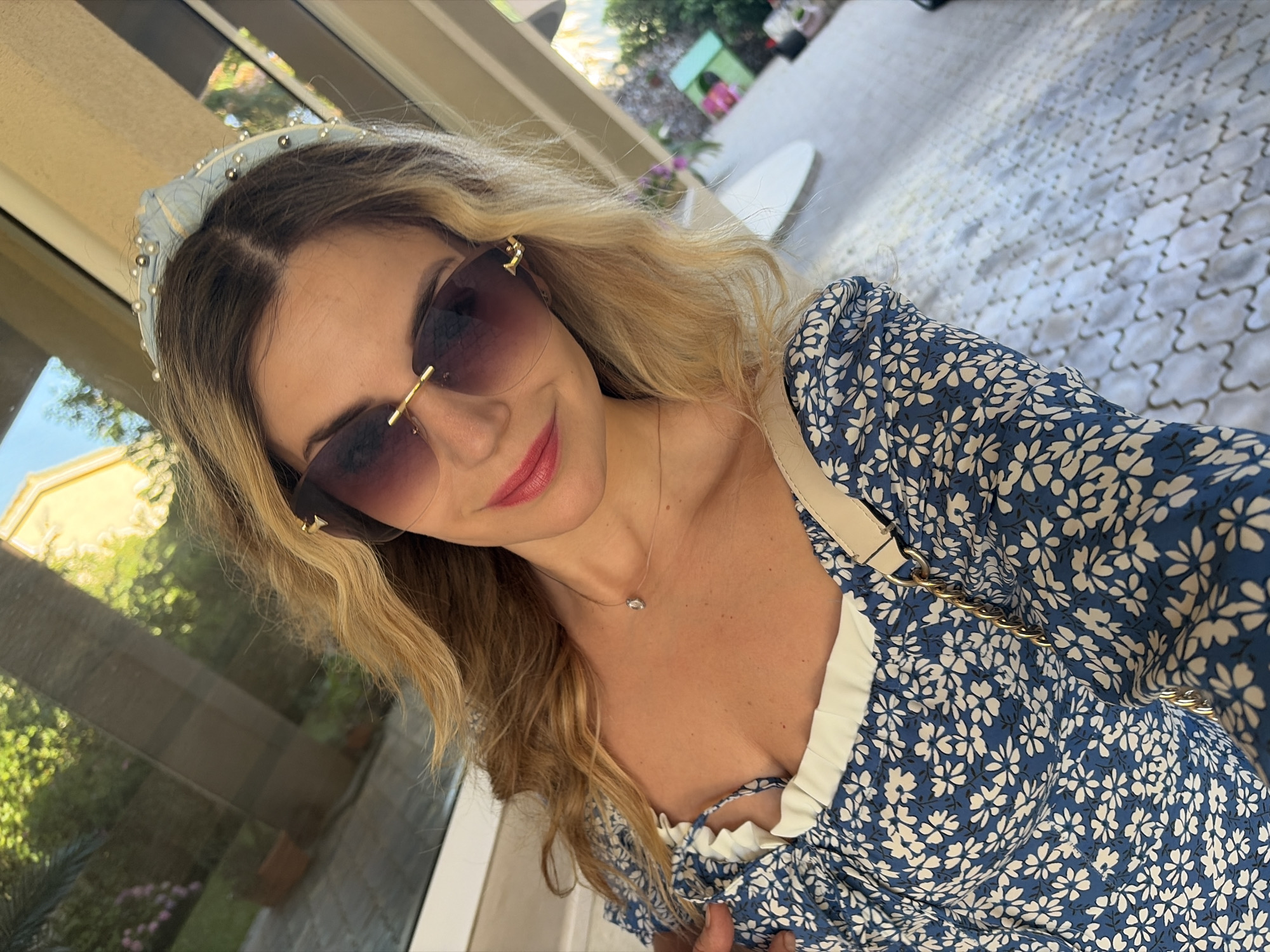 A young woman is taking a selfie outdoors. She has blonde hair and is wearing sunglasses, a blue floral dress, and a light blue headband adorned with pearls and beads. A light-colored purse with a gold chain strap is slung over her shoulder. Her chest and neck are visible, and she is wearing a delicate necklace with a single clear pendant. The background appears to be a patio or outdoor seating area with light-colored paving stones and some greenery visible through a reflective surface, possibly a window or glass door. The lighting suggests it is daytime. The image does not provide any visual cues to suggest the location is Budva, Montenegro.