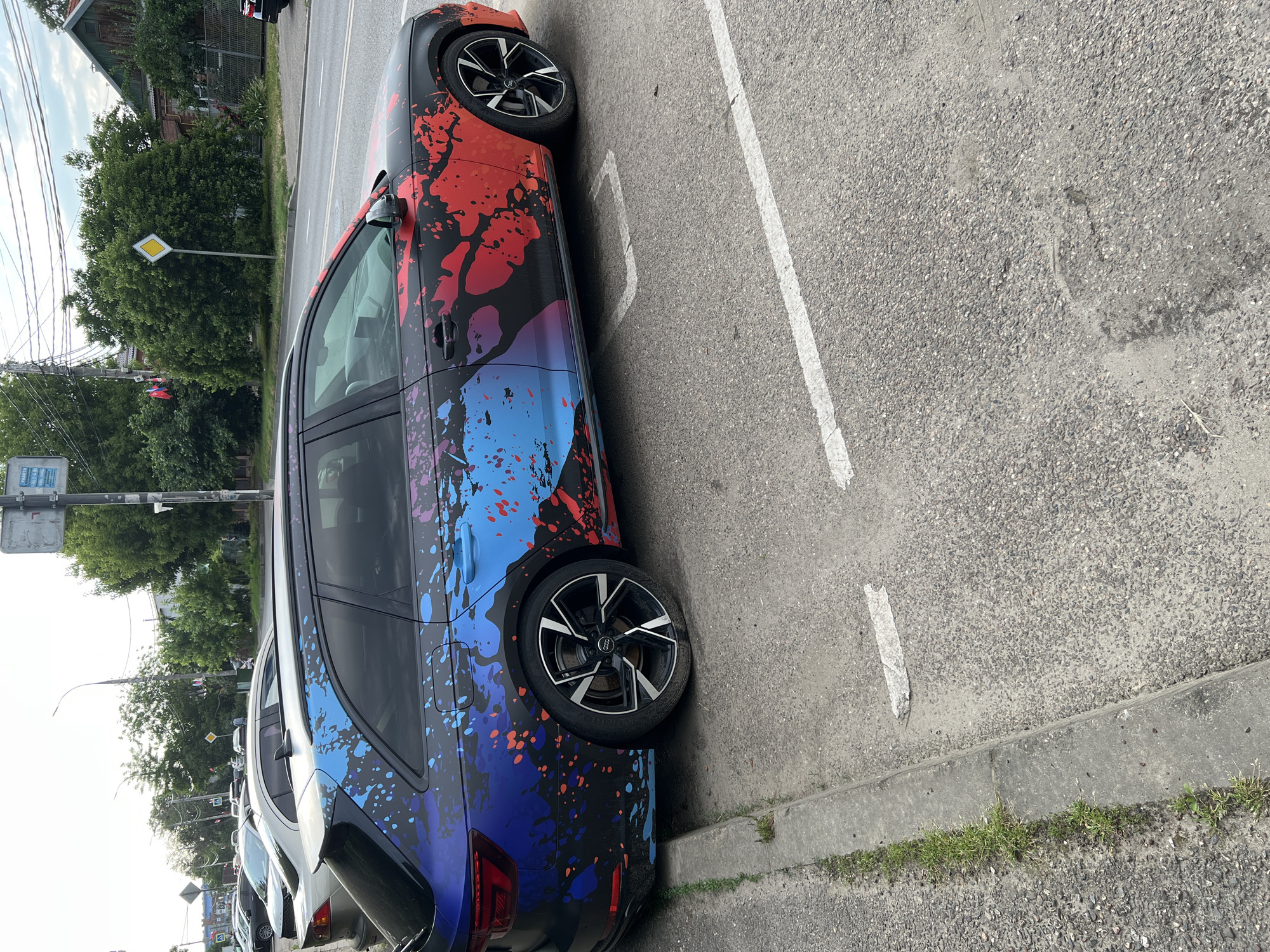 This image captures a street scene in Krasnodar, Russia. The foreground features a black Audi wagon with a distinctive abstract paint splatter design in blue, purple, and red. The car is parked on an asphalt surface, with a concrete curb and a strip of grass to its left. White painted lines demarcate parking spaces. In the background, other cars are visible parked on the street. Trees line the right side of the road, and buildings are present beyond them. A traffic sign with a yellow diamond shape is visible, along with power lines overhead. The sky is overcast. There are no people visibly present in the image.