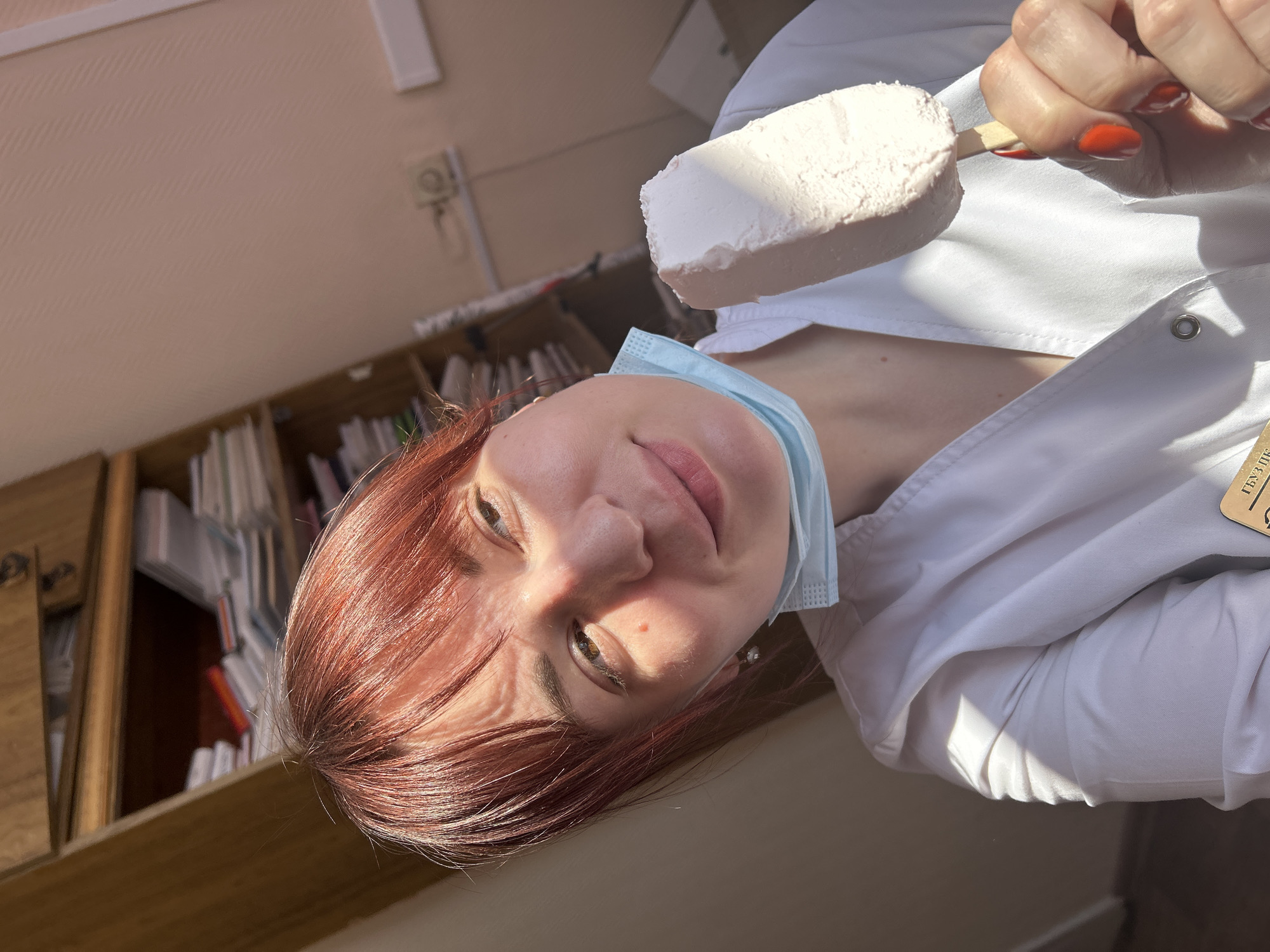 A person is holding a white ice cream bar with a wooden stick in their right hand. The person is wearing a white coat and a light blue face mask. The person has red hair. The location is in Фоки, Russia. A bookshelf with papers is in the background. The person appears to be indoors.