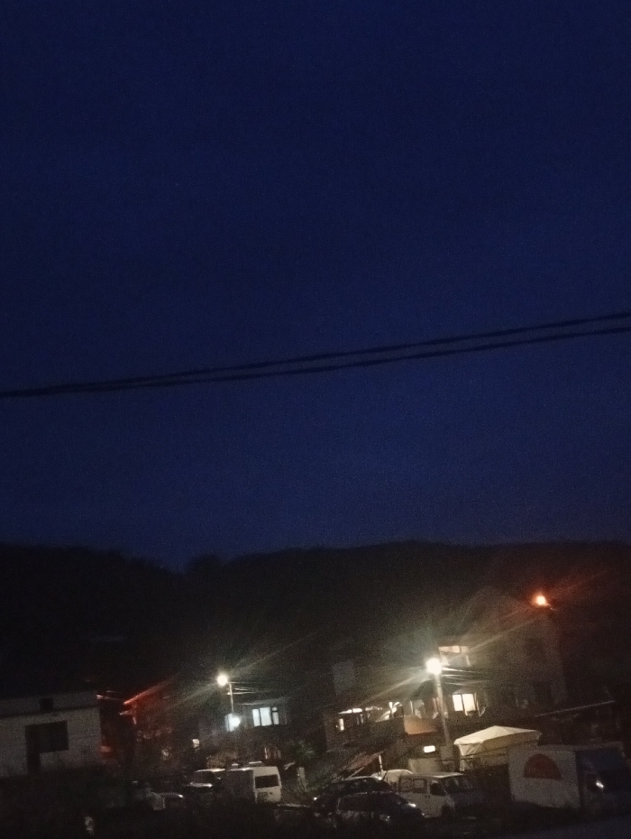 This image depicts a nighttime scene in Lebnitsa, Bulgaria. The upper half of the frame shows a dark blue sky, traversed horizontally by two dark, parallel electrical or utility lines. Below these lines, the background consists of dark, undulating hills heavily obscured by shadow, suggesting dense vegetation.
The midground reveals a residential area with multiple buildings, several of which display illuminated windows, casting a warm yellow-white light. Two prominent streetlights, one on the left and another on the right, emit strong, starburst-like glows, illuminating their immediate surroundings. A variety of parked vehicles are visible on an unpaved or dimly lit surface in front of the buildings, including several light-colored vans and darker passenger cars. A white canopy or awning is noticeable on the right side of the midground, adjacent to a vehicle. The foreground is dark and indistinct. No individuals or specific actions are discernible within the frame.