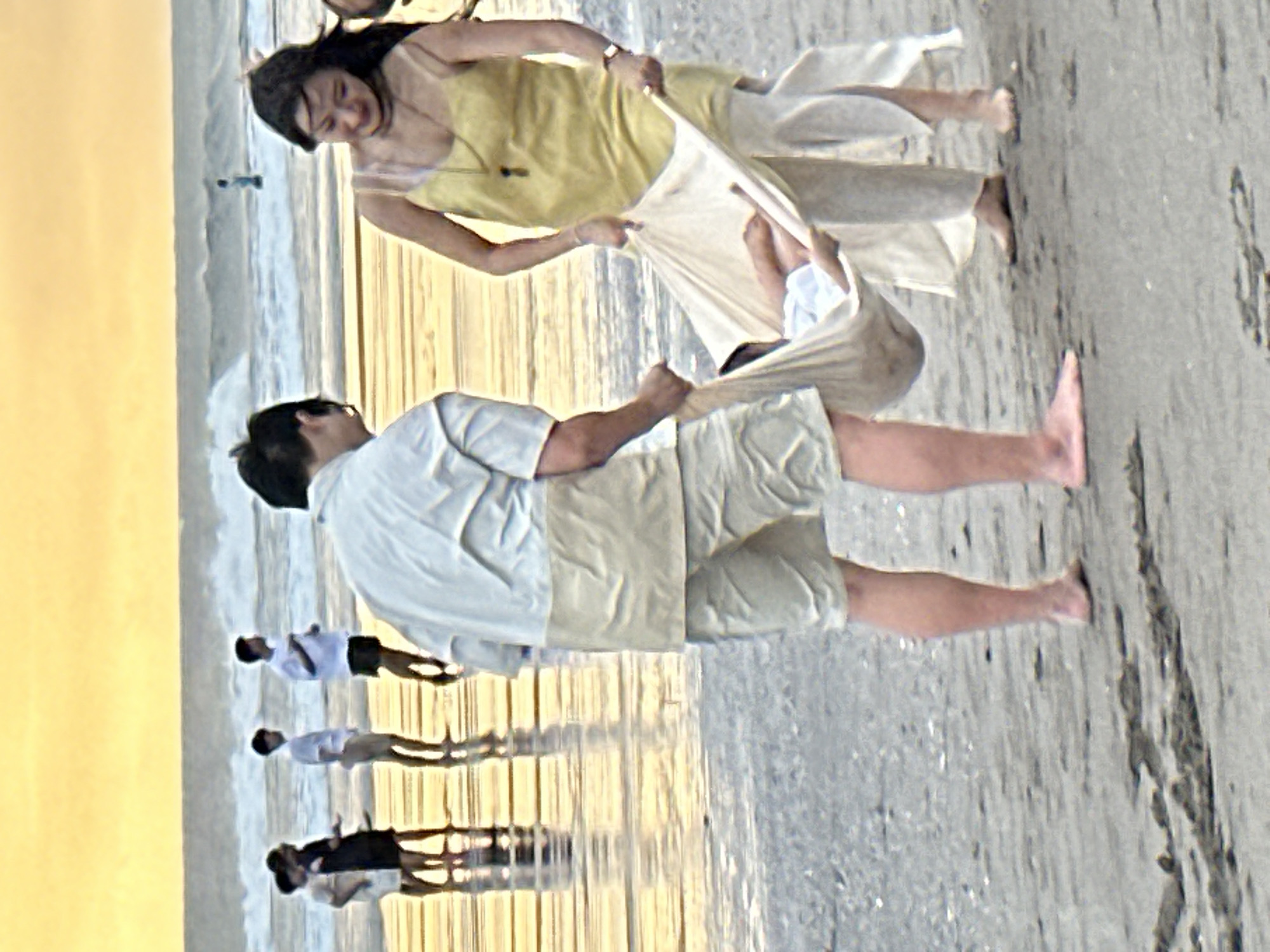 In North Kuta, Indonesia, on a beach at dusk, a couple is carrying a baby in a cloth sling. The man, wearing a light blue and tan short-sleeved shirt and tan shorts, is holding one side of the sling. The woman, in a yellow sleeveless dress and light-colored pants, is holding the other side of the sling. The baby is visible in the sling, wearing light blue and white clothing. In the background, several other people are standing on the wet sand near the ocean. The sun is low in the sky, casting a warm, yellow-orange glow over the scene and creating reflections on the wet sand. Waves are breaking in the ocean.