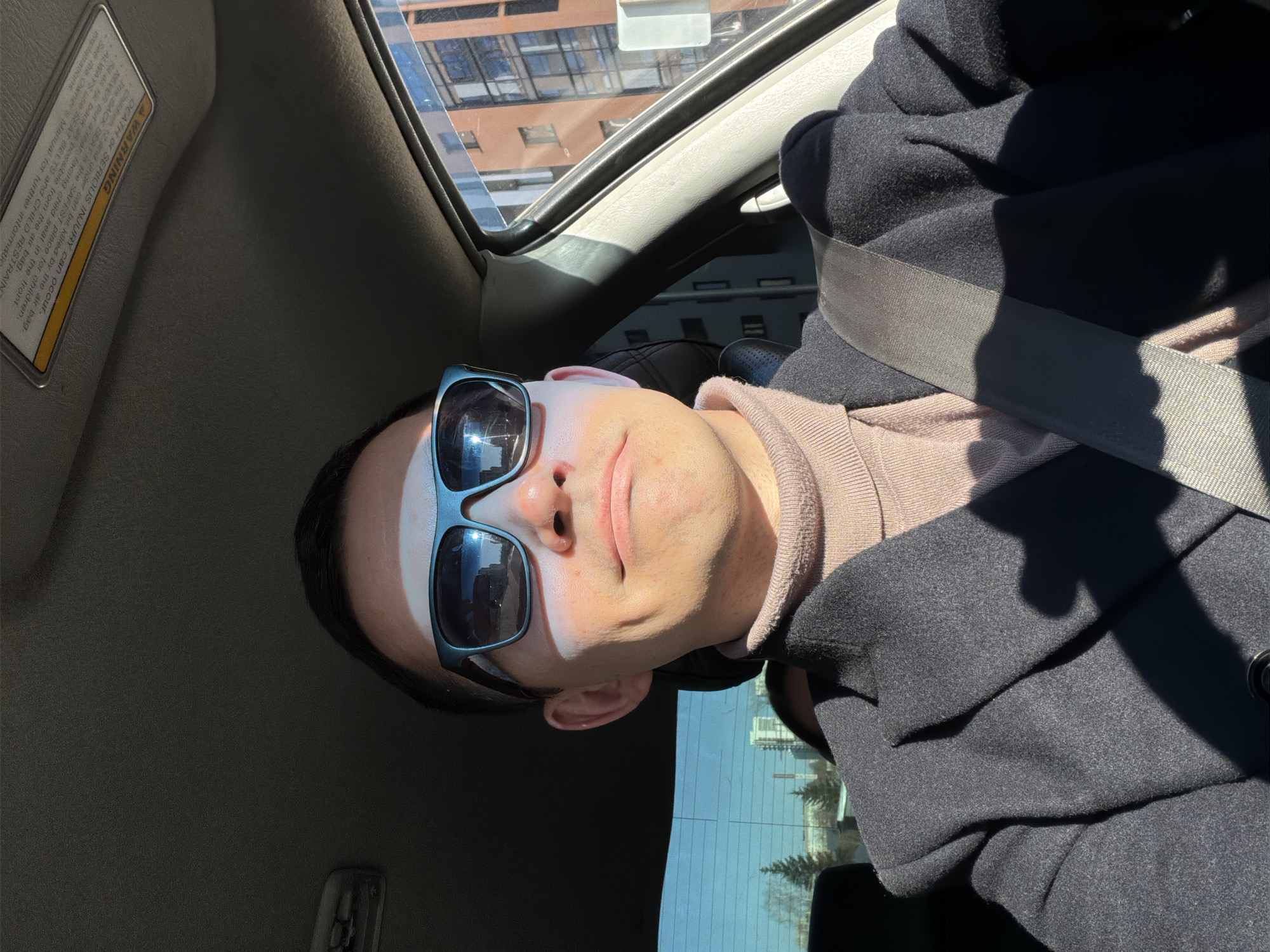 Here is a detailed description of the image:
The image shows a man in a car, wearing sunglasses and a coat. He is wearing a seatbelt. The sun is shining on his face. There is a building visible through the car window. The interior of the car is visible, including the dashboard and sun visor. The visor contains a warning label that provides instructions on child safety and passenger safety. The man is positioned in the driver's seat and may be the driver of the car. He appears to be in Izhevsk, Russia as the buildings through the car window look Russian in origin.