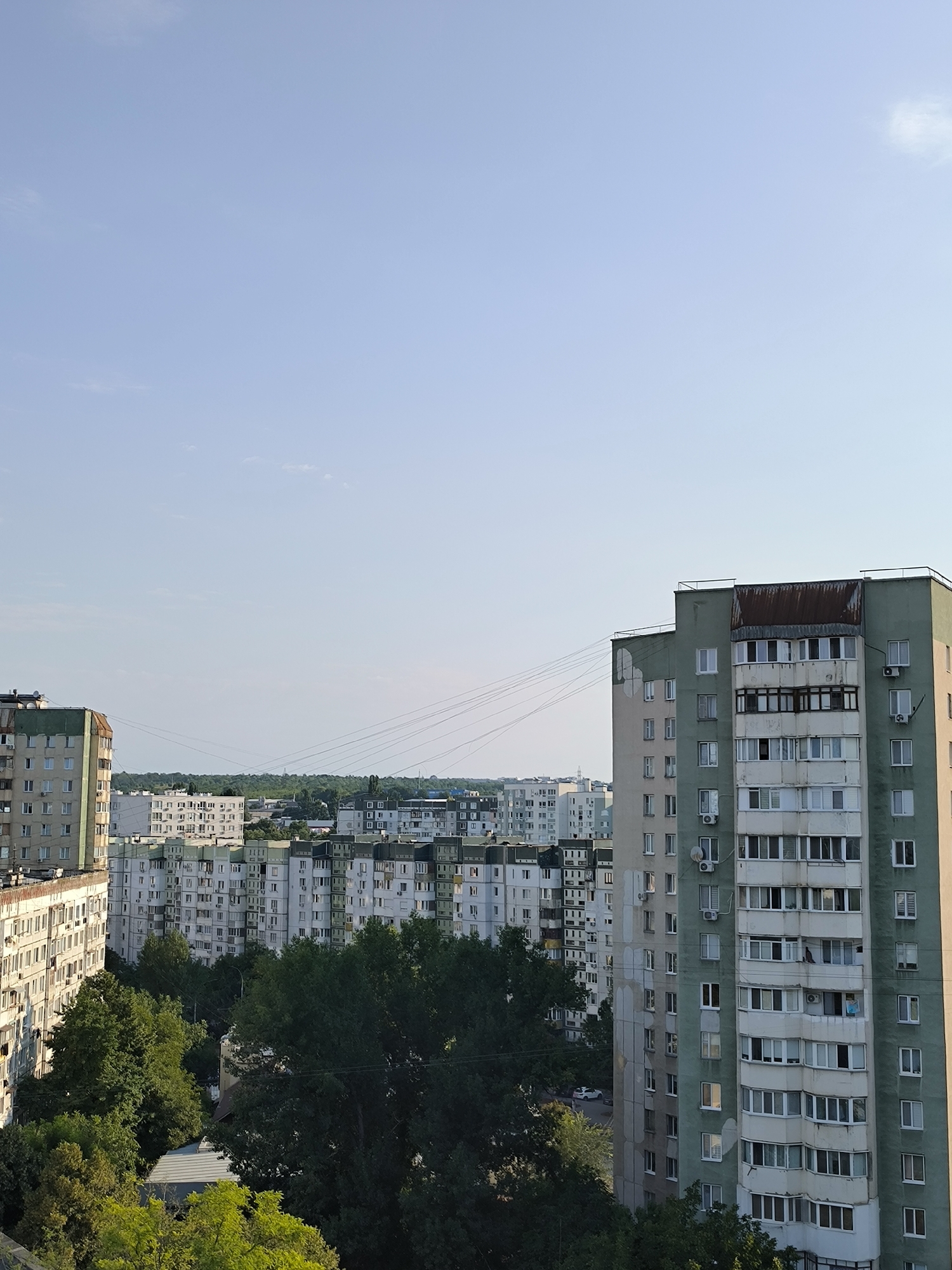 This is an outdoor, high-angle shot of a cityscape in Chișinău, Moldova. The foreground features a tall, light green residential building with numerous windows and several air conditioning units attached to its facade. The building also has a rusty, corrugated metal roof extension. To the left, another residential building, partly visible, is rendered in a pale beige color with many windows. In the mid-ground, a dense collection of similar apartment buildings, mostly white or light gray with green accents, stretches across the horizon. A line of lush green trees is situated between the foreground building and the mid-ground structures, partially obscuring some of the lower floors of the buildings. The background consists of a clear, pale blue sky with a few wispy clouds. Power lines are visible stretching across the middle of the frame, connecting distant points. The overall impression is of a densely populated urban residential area.
