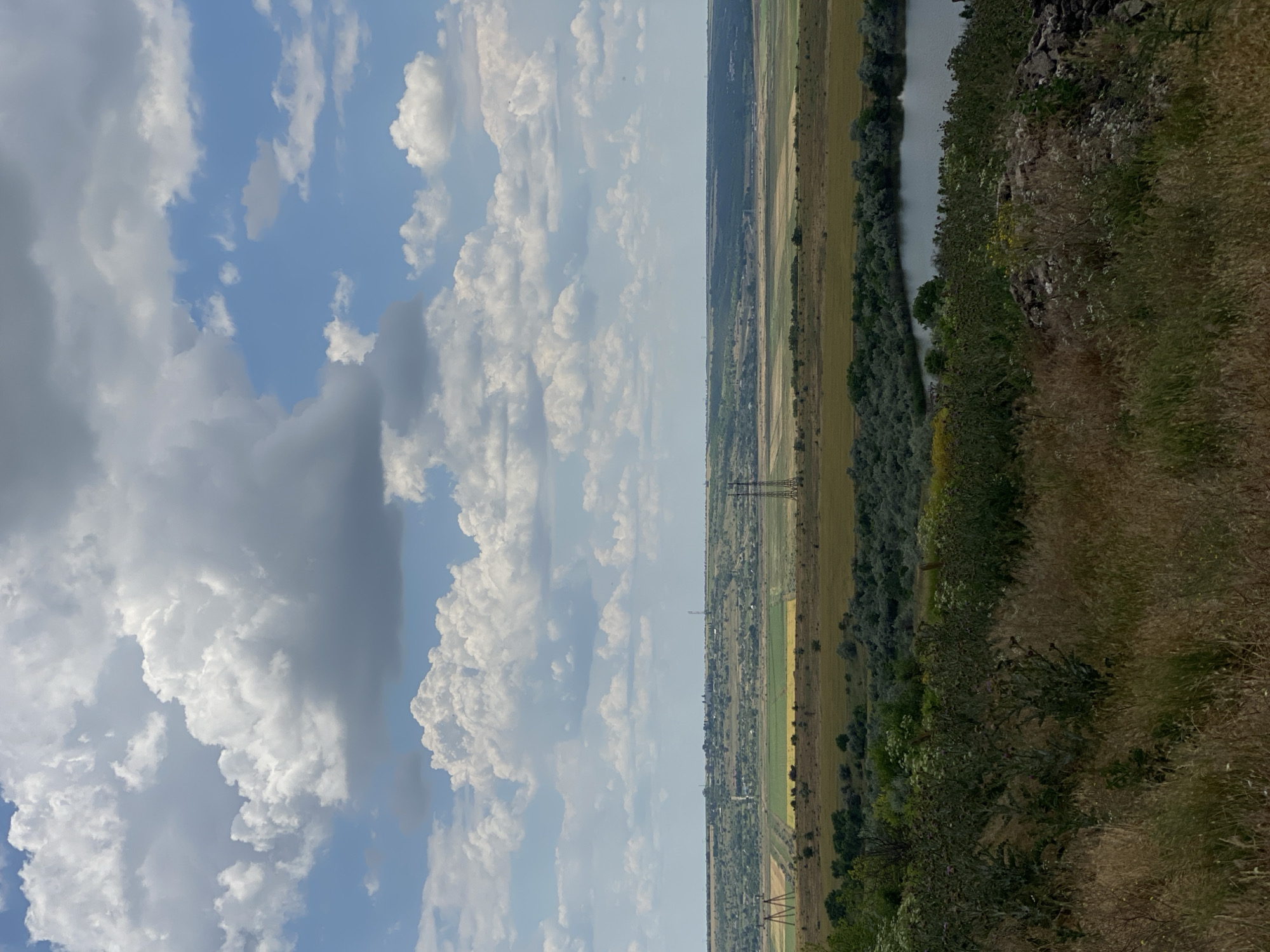 A scenic view of the rural landscape surrounding Cairaclia, Moldova, is captured on a cloudy day. In the foreground, tall, dry grass and some green bushes occupy the lower portion of the frame. A rocky outcropping is visible on the right side of the foreground. Moving into the midground, a calm body of water, possibly a lake or river, is present, bordered by dense green trees. Beyond the water, a wide expanse of fields unfolds, featuring patches of green and yellow crops, interspersed with scattered trees and shrubs. Power lines and transmission towers are visible across the landscape, indicating human infrastructure. In the far background, a distant settlement or town can be seen nestled on a gently rolling horizon, with more fields and vegetation stretching towards it. The sky above is dramatic, filled with a mix of large, fluffy white and grey clouds, with patches of blue sky peeking through. The overall impression is one of a vast, agricultural region under a dynamic sky.