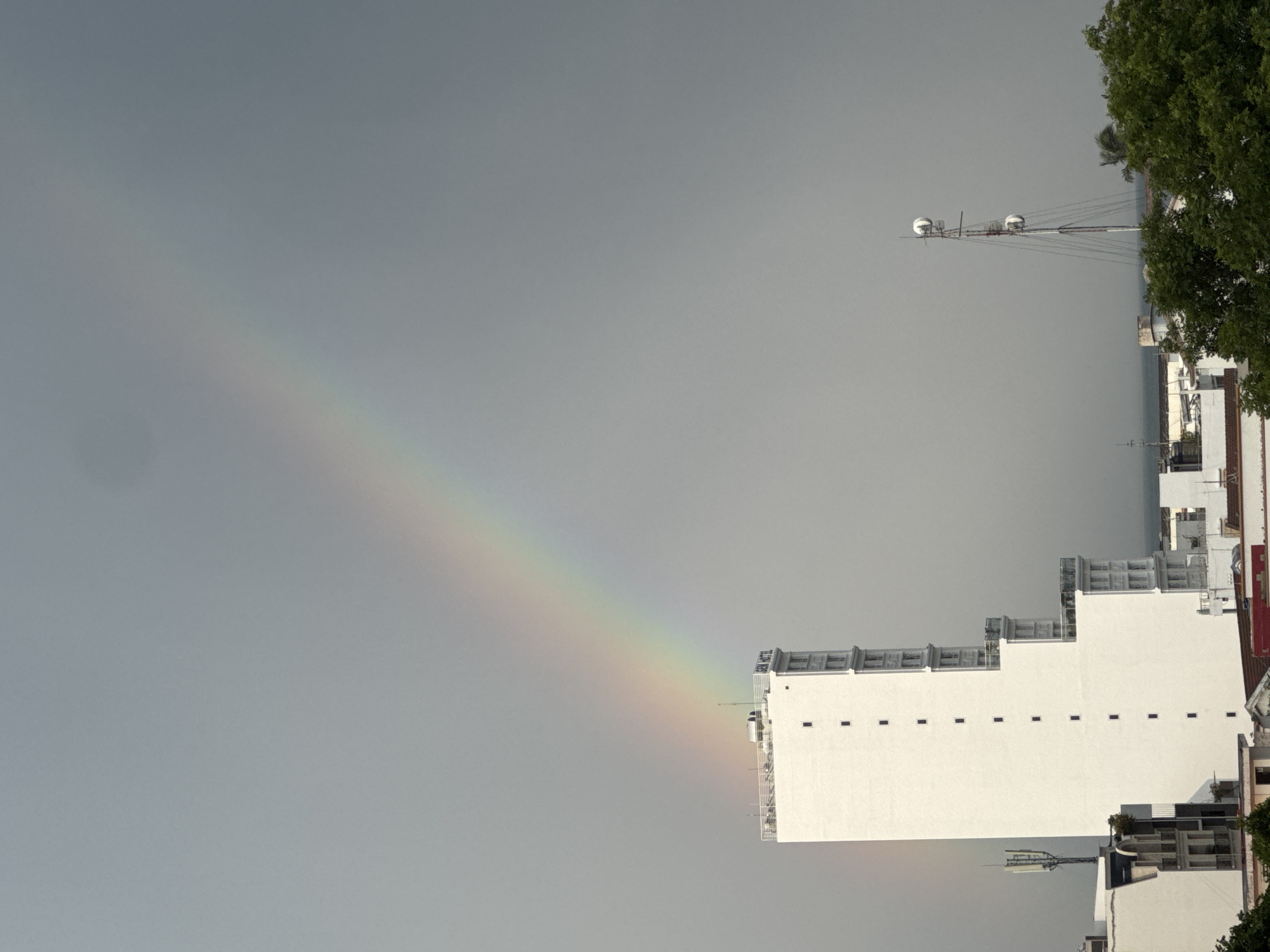 This image, captured in Nha Trang, Vietnam, depicts a city skyline under a gray, overcast sky, with a rainbow arcing across the upper portion of the frame. The most prominent structure is a tall, white, rectangular building with a flat roof and a railing. Several small, dark rectangular windows are visible on its facade. The building has a nameplate on its upper right side that reads "GEN HOTEL". To the left of this building, a smaller, white building is partially visible, with an antenna tower rising from its roof. To the right of the main building, a tall, slender communication tower with multiple antennas is erected. The lower portion of the image reveals the rooftops of other buildings and lush green trees. The sea is visible in the distance, beyond the city structures. The overall scene is of an urban environment, likely captured after a rain shower, given the presence of the rainbow.