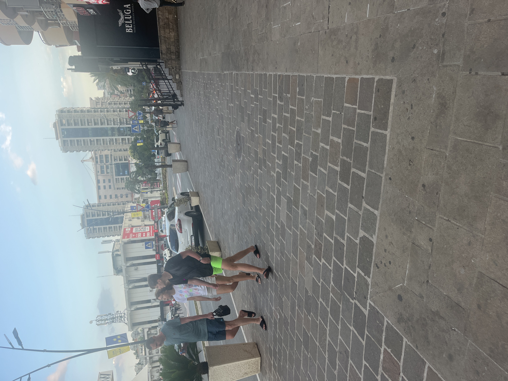 The image shows a street scene with several people walking on a paved sidewalk. In the background, there are tall buildings, palm trees, and cars. The sky is visible with some clouds. The time of day appears to be daytime, and the weather seems to be clear.
The subjects in the foreground are three people walking from left to right. The person on the left is wearing a dark t-shirt and blue shorts. They are holding a dark object, possibly a phone, in their left hand and a dark hat in their right hand. The person in the middle is wearing a white t-shirt with a colorful design and light-colored shorts. The person on the right is wearing a black t-shirt and bright green shorts. They are all casually dressed, suggesting a relaxed atmosphere.
The scene is a city street, with a modern urban feel. The sidewalk is made of gray cobblestones. Behind the people, there is a road with several cars, including a white car with "TAXI" visible on it. Some of the buildings in the background have signage, including one that says "BELUGA NOBLE RUSSIAN VODKA". There are also traffic signs visible. The overall impression is a busy urban environment on a pleasant day. There are no strong emotional cues visible, but the casual attire and relaxed posture of the people suggest a normal, everyday occurrence.